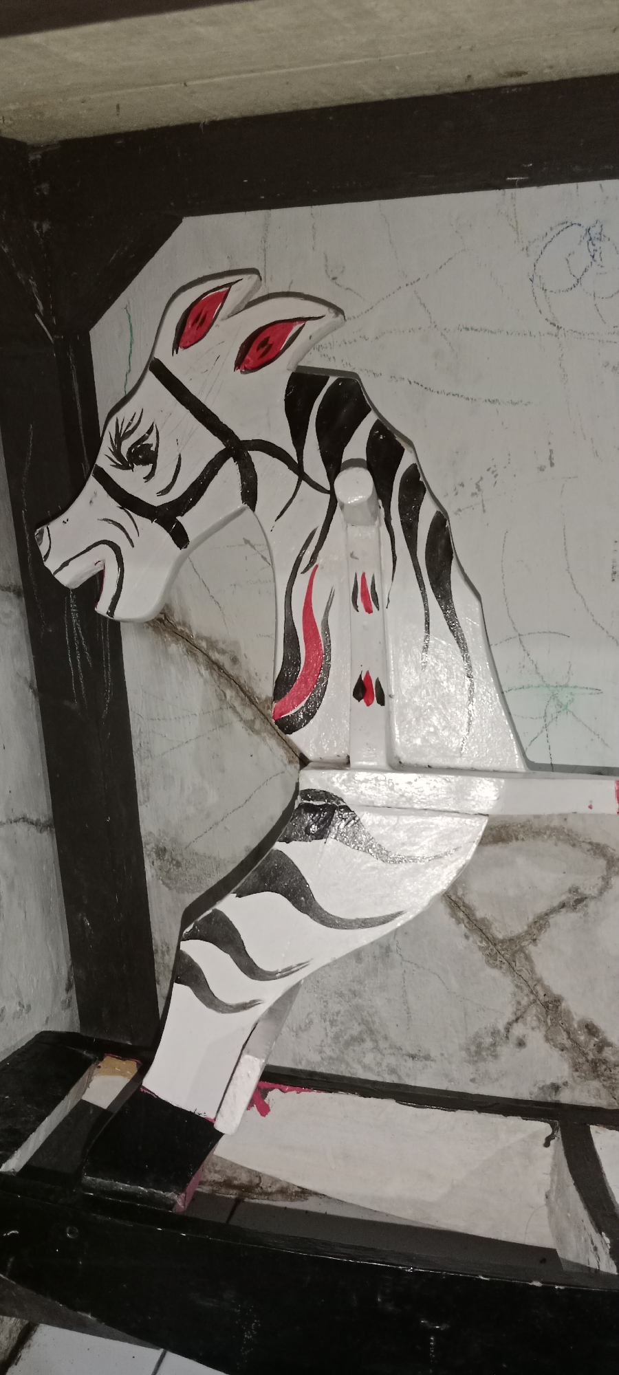 This is a close-up, eye-level, indoor shot of a stylized, white wooden horse head with black and red markings, likely part of a larger sculpture or decorative piece. The horse head is positioned facing left. It has red eyes outlined in black, and black stripes that mimic a mane and bridle. The surface appears to be painted wood, and there are some faint scratches and smudges on the wall behind it, which is also painted. The lighting suggests an artificial light source. The image is cropped, so the full context of the object and its surroundings is not visible. The location is a workshop or storage area in Subang, Indonesia.