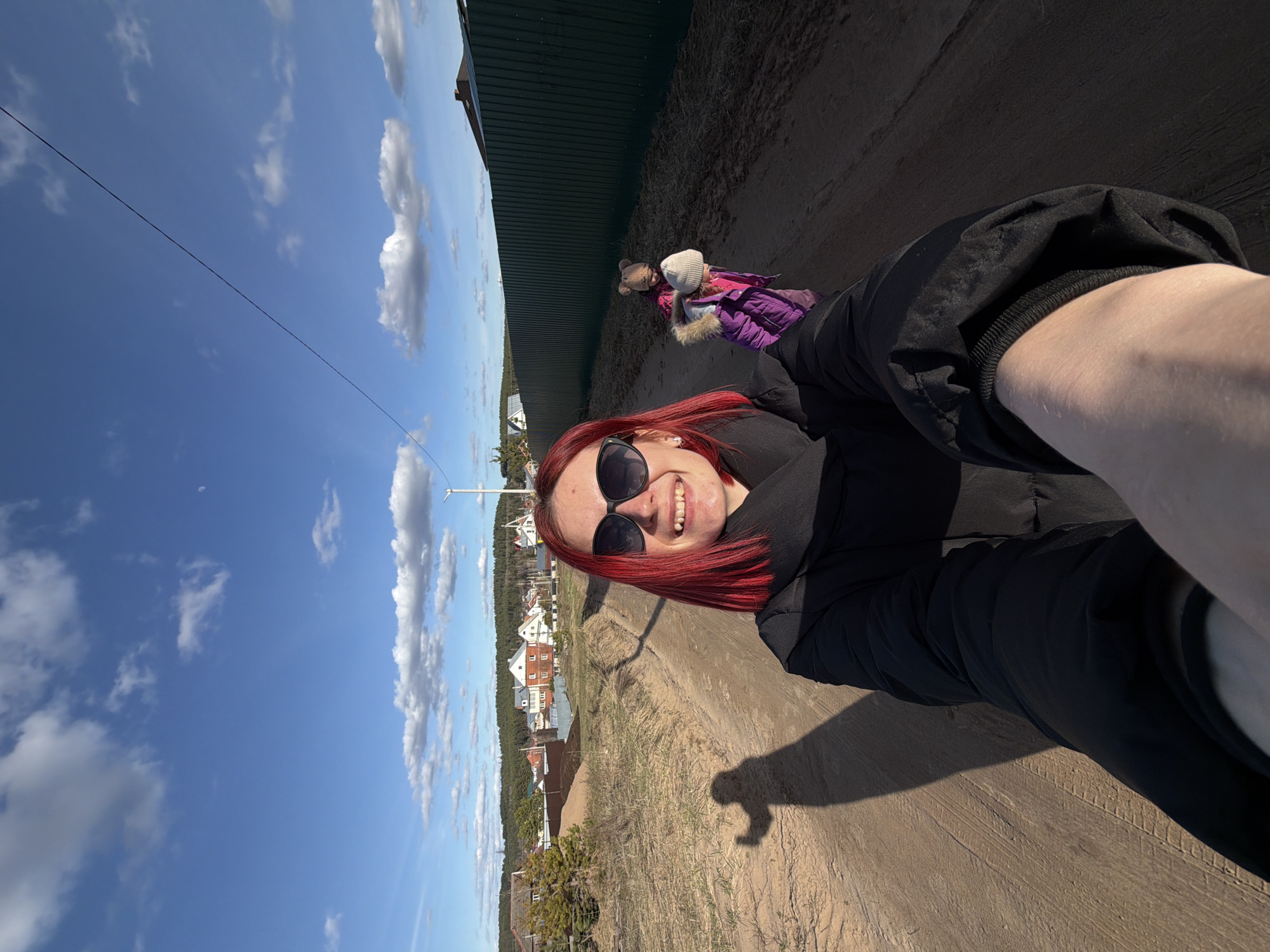 A selfie shows a woman with red hair and sunglasses standing on a sandy path. Behind her is a green fence, a child lying on the ground with a teddy bear, and houses and trees in the distance under a blue sky with clouds. The location is Chaikovsky, Russia.