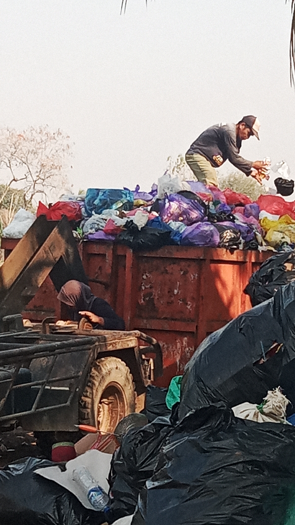 This is a vertical, medium shot of a refuse collection scene in Sungai Sipai, Indonesia. A large, rusty, orange dumpster overflows with colorful plastic bags filled with what appears to be clothing and other discarded items. Two individuals are visible. In the upper right, a man wearing a baseball cap and a dark long-sleeved shirt is reaching into the dumpster, sorting through the trash. In the lower left, a person wearing a dark headscarf and dark clothing is bent over in the dumpster, also engaged with the contents. The background is a pale, hazy sky with the bare branches of a tree visible on the left. In the foreground, several large black plastic trash bags are piled up, obscuring parts of the dumpster and a vehicle with a prominent tire.