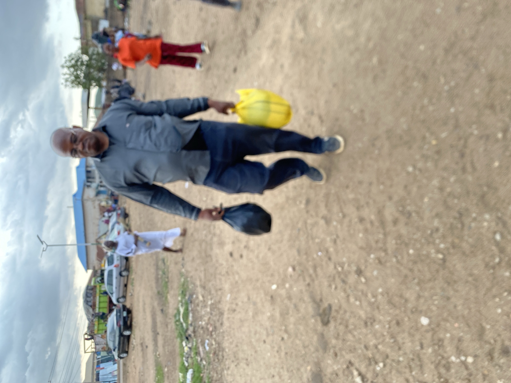 An individual is walking on a dirt and gravel path in Bukuru, Nigeria. The person has dark skin, a shaved head, and is wearing a grey button-down shirt, blue pants, and blue sneakers. They are holding a yellow bag in one hand and a dark-colored hat or bag in the other. Other individuals are visible in the background, one wearing orange and red clothing and another wearing white clothing. There are several vehicles parked along the side of the path, and there is a blue-roofed building in the background. The sky is cloudy. Trash and vegetation are visible along the path.