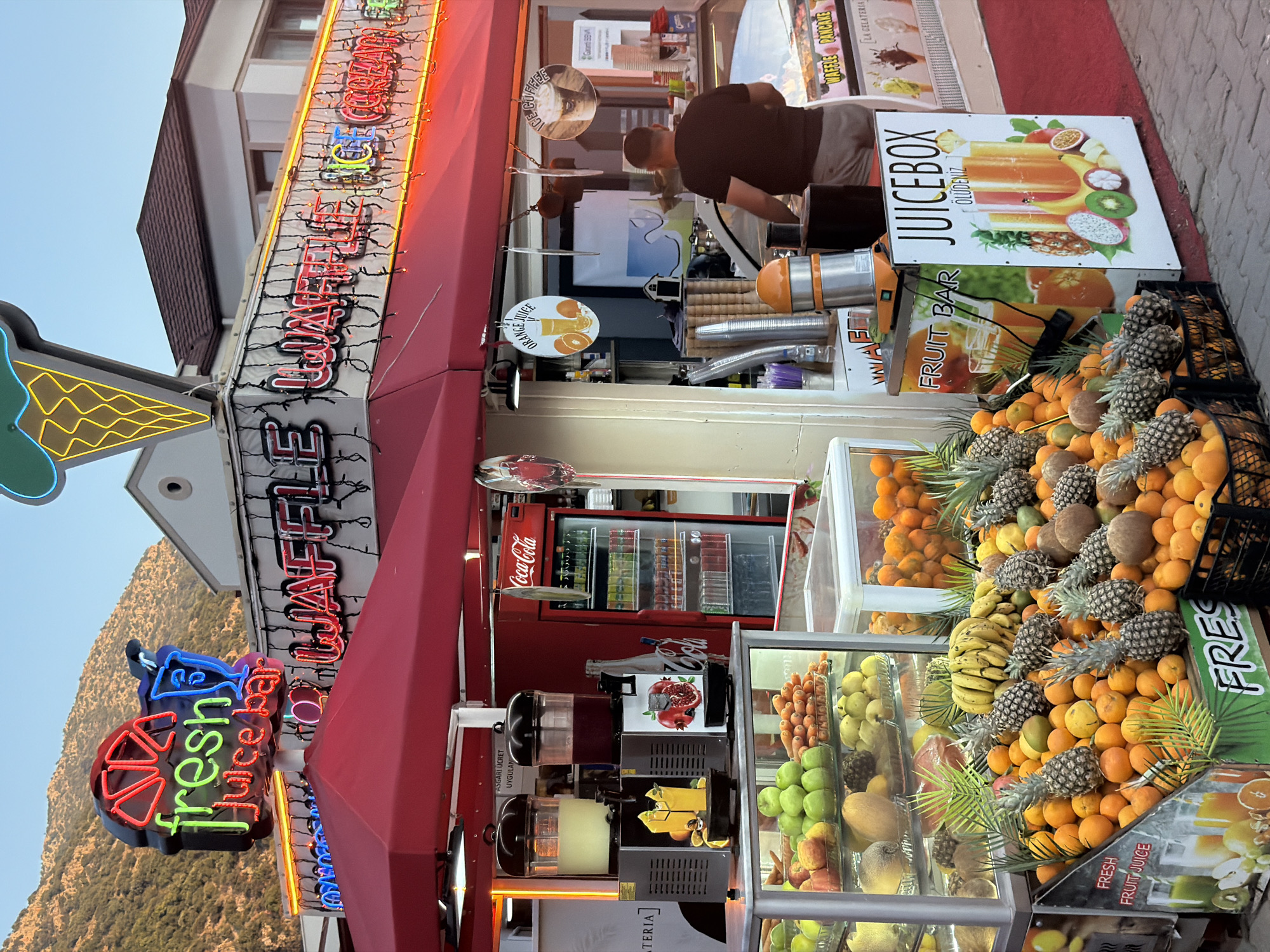 The image displays a bustling outdoor juice and waffle bar, captured likely during the evening with its neon lights fully illuminated. The establishment is set against a natural backdrop of a large mountain, hinting at a scenic location like Fethiye, Turkey.
The stall boasts an impressive and colorful array of fresh fruits, including abundant oranges, pineapples, bananas, coconuts, apples, and various melons, neatly arranged in display cases and crates. Several juicing machines and a Coca-Cola refrigerator are visible behind the counter. A person in a dark t-shirt is seen from behind, preparing items at the counter.
The bar is adorned with numerous signs and neon lights. Prominent neon signs spell out "fresh Juice bar" and "Waffle Pancake." Other illuminated signs advertise "ICE COFFEE" and "ORANGE JUICE." A large, eye-catching sign reads "JUICEBOX ÖLÜDENİZ," featuring an enticing illustration of a juice glass filled with tropical fruits. Further signs include "FRUIT BAR," "FRESH FRUIT JUICE," and a Turkish sign stating "ASGARİ ÜCRET UYGULANIR" (minimum wage applied). The visible text "LA GELATERIA" suggests ice cream is also available.
The combination of vibrant neon lights, the fresh fruit display, and the visible activity creates an inviting and lively atmosphere, characteristic of a popular tourist spot offering refreshing drinks and sweet treats.