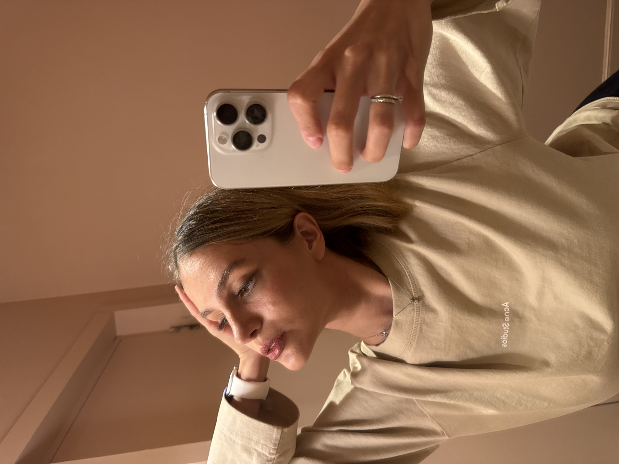 A woman is holding a white smartphone up to the mirror, taking a selfie. She has short blonde hair and is wearing a light beige, long-sleeved shirt with "Acne Studios" printed on the right side of the shirt. She is wearing a ring on her left ring finger and a white smartwatch on her left wrist. The background appears to be a room with light beige walls, and the photo is taken indoors. The location is unknown and cannot be discerned from this image alone. The city of Tbilisi, Georgia cannot be discerned.