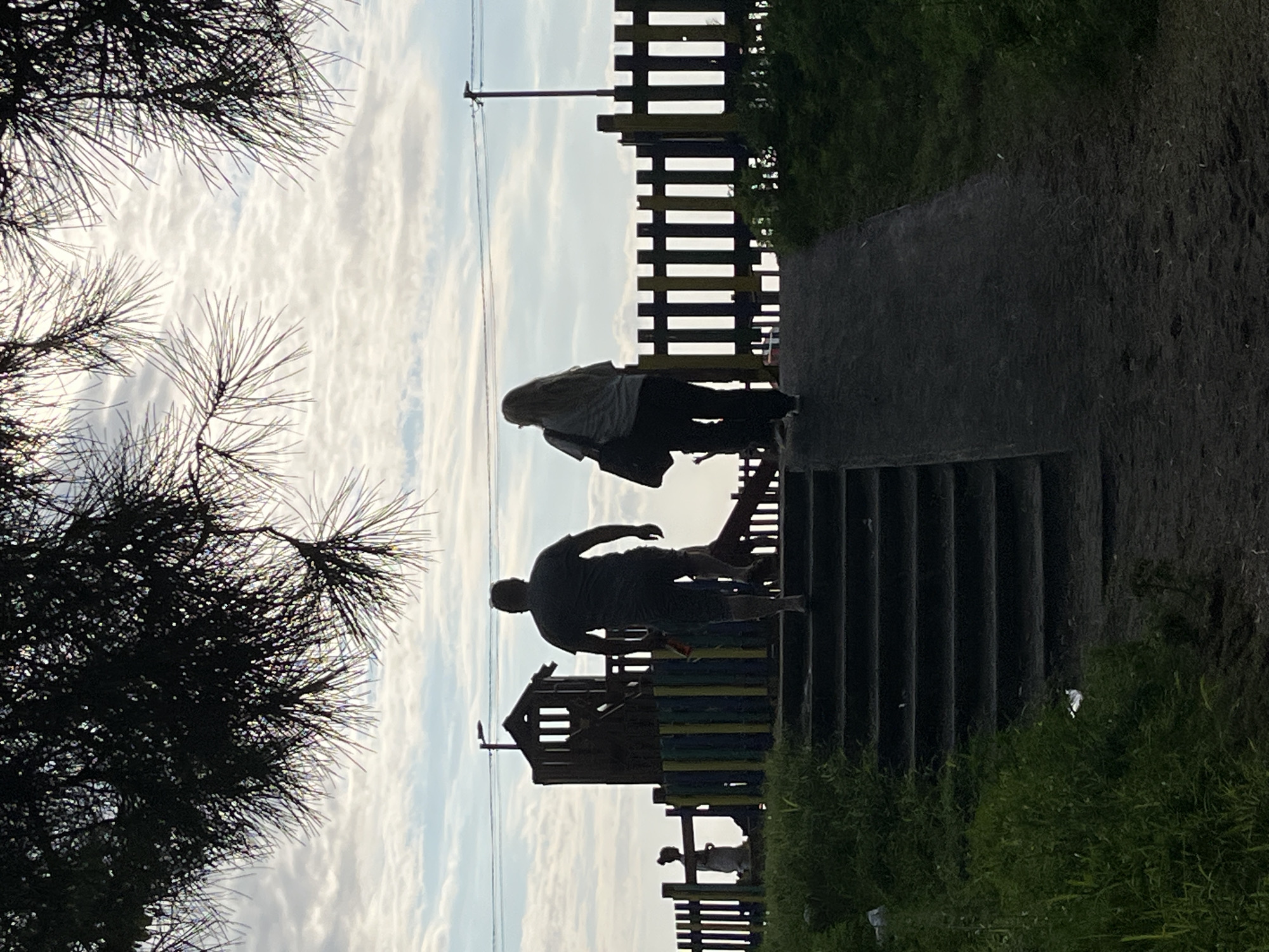 In Ureki, Georgia, at dusk, a man and a woman walk up a ramp next to a set of stairs, heading towards a playground. The man, in silhouette, wears a dark t-shirt and shorts, with a thin red object clutched in his right hand. The woman, also in silhouette, carries a dark bag over her left shoulder and wears glasses. Behind them, the playground features a wooden structure and a colorful fence. The sky is cloudy, with a hint of the setting sun. Green grass and foliage line the path.