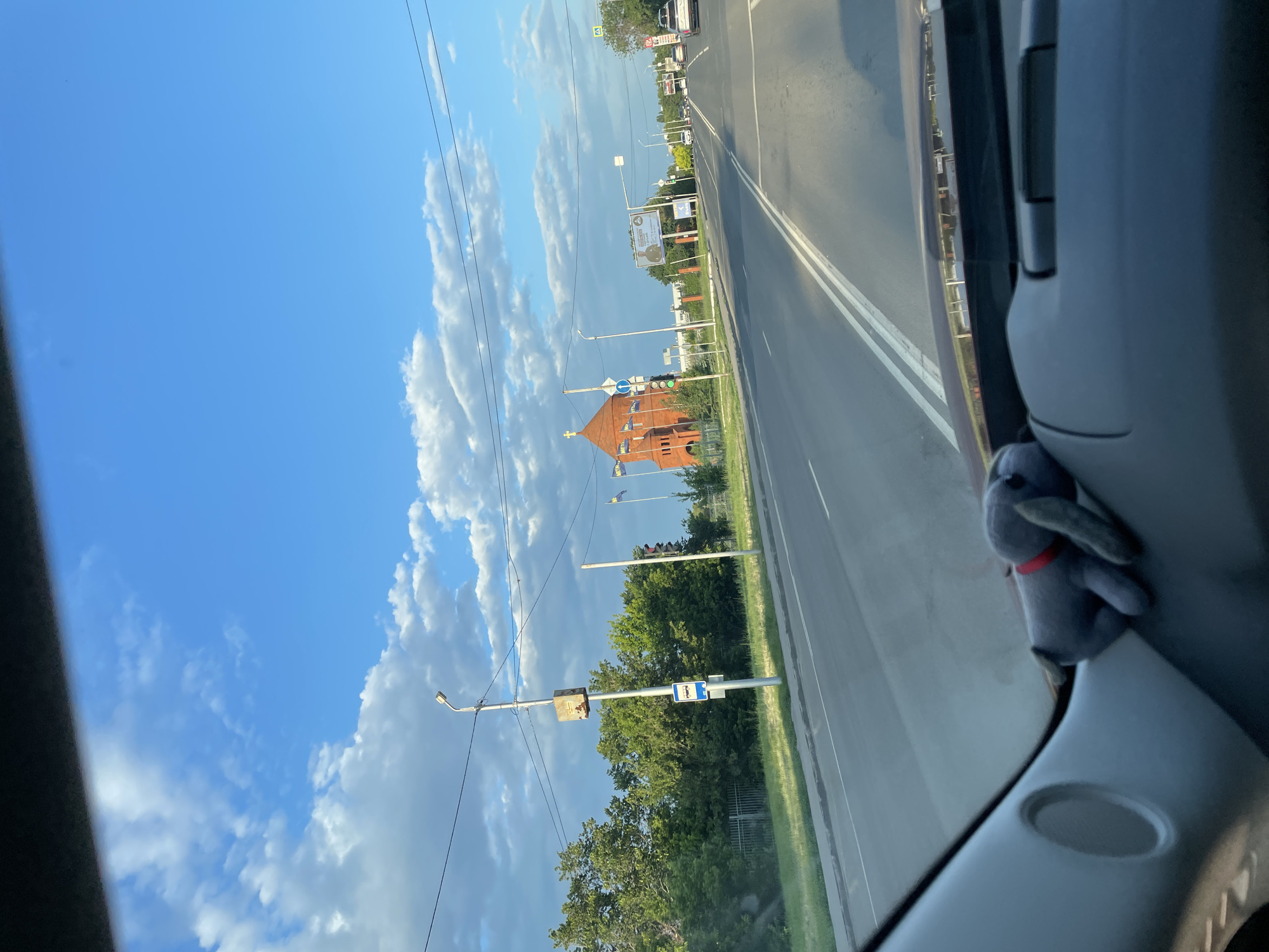 The image shows a street view from inside a car, with a plush toy dog on the dashboard. The road is paved with lane markings, and there are trees and buildings on either side. A prominent red brick church with a cross on top is visible in the distance, along with several flags. Traffic signals and streetlights are also present. The sky is blue with scattered clouds. Based on the architecture of the church and the presence of specific flags, this scene is likely located in Saratov, Russia, potentially near the Church of the Resurrection of Christ, given its distinctive appearance and common presence in this region. The time of day appears to be late afternoon or early evening, judging by the lighting.