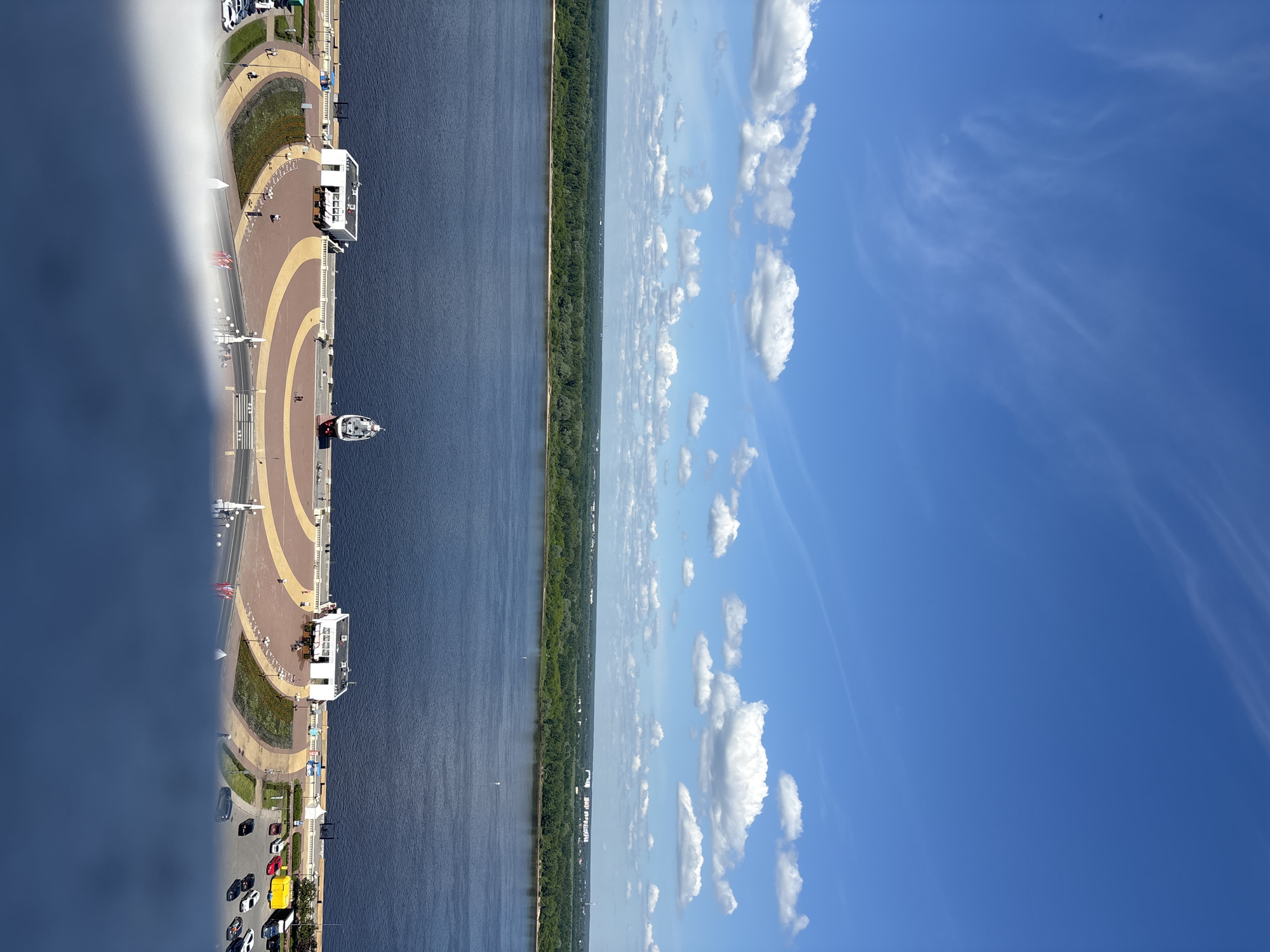 This aerial or high-angle shot captures a scenic riverfront in Nizhny Novgorod, Russia, under a bright, clear blue sky dotted with scattered white cumulus and wispy cirrus clouds. The wide, dark blue river forms a prominent horizontal band across the middle of the frame.
On the far bank, a continuous line of dense green forest stretches along the horizon. The near bank features a well-developed promenade with distinctive light brown and yellow patterned paving, curving gently alongside the water. Interspersed green landscaped areas and flower beds add color to the embankment.
Several individuals can be seen walking leisurely on the promenade, enjoying the day. Prominent white buildings, likely cafes or visitor centers, with outdoor seating areas, are situated along the waterfront. A boat, appearing to be a small passenger vessel or yacht, is visible on the river near the promenade. Further inland, a road runs parallel to the river, featuring several parked or moving vehicles, including a noticeable yellow truck, a red car, and various dark-colored cars. Streetlights line the promenade.
The image conveys a pleasant and peaceful atmosphere on a clear, sunny day, likely during midday or early afternoon. The bright conditions and activity suggest a popular recreational area. No discernible text is clearly legible on any objects, buildings, or signs within the image.