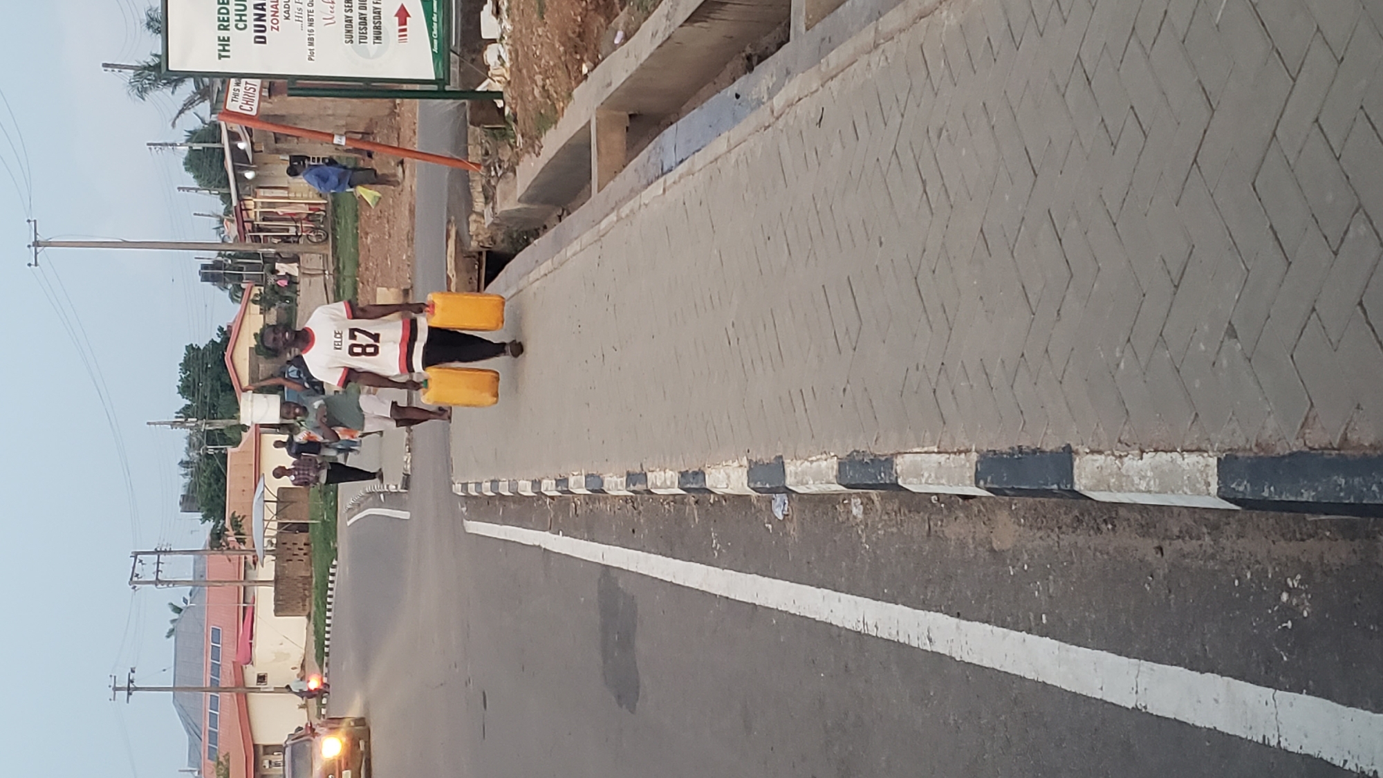 The image depicts a roadside scene in Narayi, Nigeria, likely during dusk or early morning hours, indicated by the soft, diffused light. The primary subject is a man in the foreground, wearing a white jersey with "KELCE 82" printed on it. He is smiling while carrying two large yellow jerrycans, one in each hand, suggesting he is transporting water or another liquid.
He is walking on the side of an asphalt road, which features a distinct white lane marking. To his right, there's a gray paved sidewalk laid in a herringbone pattern, separated from the road by curbs painted black and white. Beyond the sidewalk, a concrete drainage ditch is visible, followed by various buildings, utility poles, and overhead power lines.
In the mid-ground and background, several other people are present, some appearing to walk. A vehicle with its headlights illuminated can be seen further down the road. On the right side of the image, a green and white sign is prominently displayed, bearing text related to "THE REDEEMED CHURCH OF GOD ZONAL HQTRS" located in "DUNA, KADUNA." The sign also lists service times, including "SUNDAY SERVICE," "TUESDAY DIGGING DEEP," and "THURSDAY FAITH CLINIC," concluding with "Jesus Christ the same..." The scene captures a moment of daily life and activity in the community.