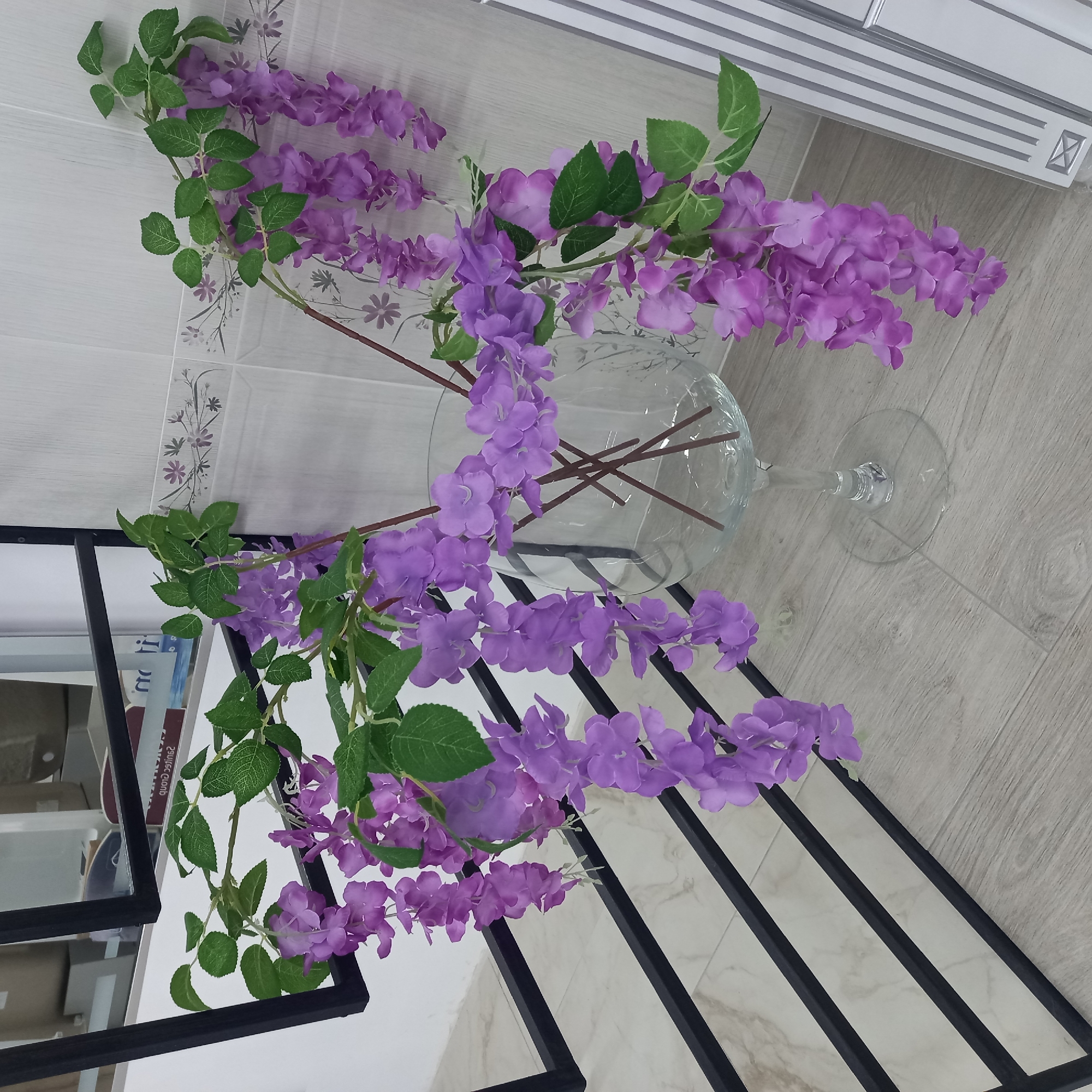 A still life photograph features a display of artificial purple flowers with green leaves arranged in a clear glass vase. The vase is positioned on a surface with a wooden grain pattern, against a backdrop of white tiled walls with a decorative floral border. The flowers cascade from the vase, with some branches extending over the edge. In the foreground and to the left, a black metal shelving unit with multiple levels is visible. The shelves are empty, except for a few books on the second shelf from the top, one of which has visible text that appears to be in Cyrillic script. The lighting suggests an indoor setting, possibly a room or a shop. The overall impression is one of simple, decorative arrangement. No people or animals are present in the image. The scene does not provide specific cues about the time of day or weather.
