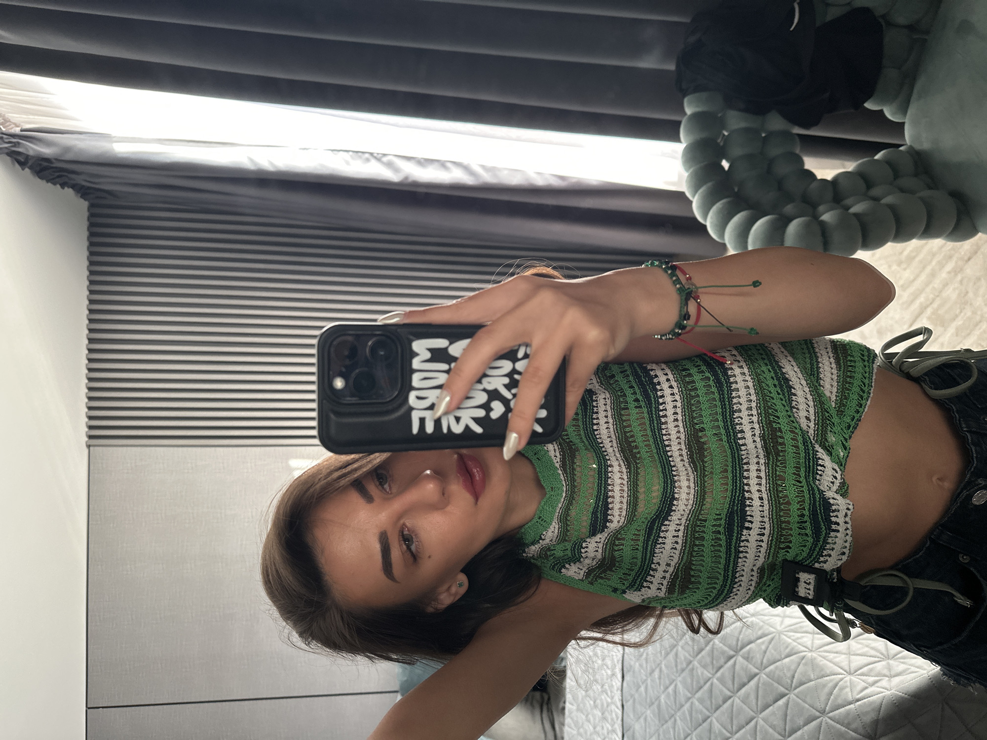 A young woman is taking a mirror selfie in a bedroom. She is wearing a green and white striped crop top and dark denim shorts. She has long brown hair and is wearing makeup. She is holding a black phone in her right hand, which is displaying text. She is also wearing bracelets on her left wrist. The background of the image shows a bed, curtains, and a chair. The room appears to be well-lit. The location is a bedroom in Sofia, Bulgaria.