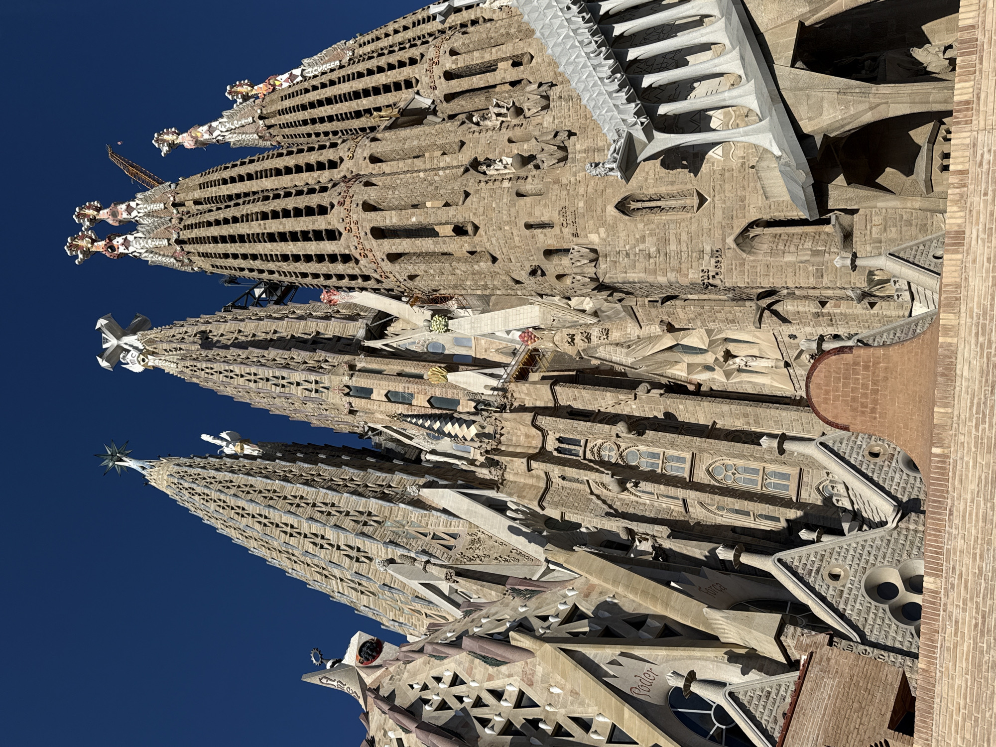 This low-angle, eye-level photograph captures a detailed view of the Sagrada Familia basilica in Barcelona, Spain, under a clear, deep blue sky. The massive stone structure dominates the frame, showcasing multiple intricately designed spires and facades.
On the left, a section of the basilica features elaborate, organic stonework with triangular cutouts and an inscription that reads "Poder" (power). A tall, ornate spire rises from this section, topped with a star-like finial. Adjacent spires further right exhibit complex patterns of window-like openings, some crowned with decorative mosaic elements resembling fruit, while another is topped with a distinct X-shaped cross. A yellow construction crane is visible between two of the central spires, indicating ongoing work.
Towards the right, another section of the façade displays more angular and blocky stonework, with vertically aligned windows and niches containing sculptures. Inscriptions like "Sanctus" (holy) and "Corda Surum" (lift up your hearts) are visible on the stone. Architectural elements include arched doorways and columns. Strong sunlight illuminates the structure from the right, creating sharp contrasts and casting deep shadows that highlight the three-dimensional complexity of the gothic and art nouveau details.
In the immediate foreground, a low, light-colored brick wall extends across the bottom of the image, with sloped, stone-tiled rooflines and small, decorative chimney-like structures partially visible above it. No individuals are present in the visible scene.