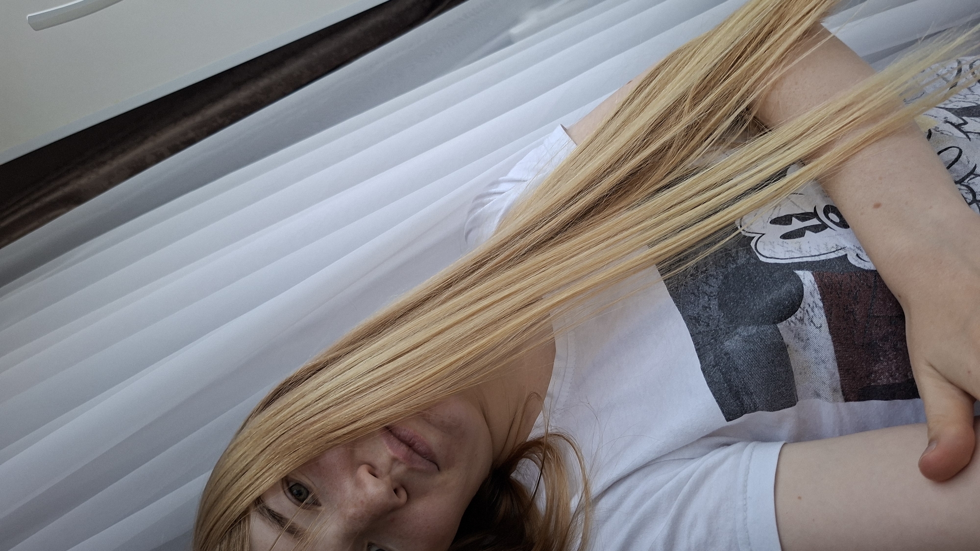 A close-up shot shows a young Caucasian woman with long blonde hair, partially obscuring her face. She is wearing a white t-shirt with a graphic print of Mickey Mouse. The background consists of white pleated blinds, suggesting an indoor setting. A dark brown stripe runs horizontally across the top of the image, above the blinds. The lighting appears bright, possibly indicating daylight. The woman's expression is neutral. The image is oriented sideways, with the woman's head on the right and her body extending towards the left.