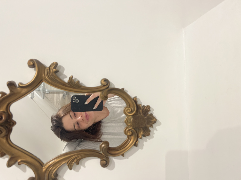 A woman with shoulder-length brunette hair holds a black smartphone, taking a selfie reflected in an ornate gold-framed mirror. She wears a light gray long-sleeved shirt. The mirror hangs on a white wall, with a glimpse of a showerhead and tiled wall visible in the reflection. The camera captures the woman's face at a slight angle, revealing a soft smile.