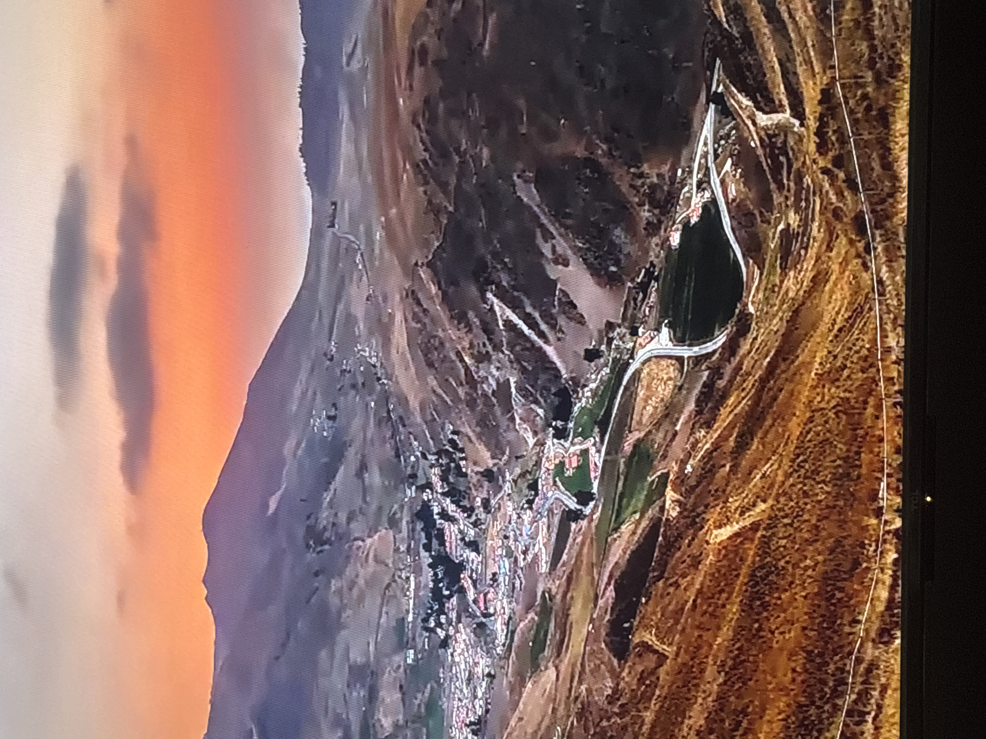 This media file is an aerial view of a landscape, possibly taken from an aircraft or a drone, with a sunset creating a vibrant orange and pink sky on the left side of the image. The sky transitions to a deep blue or purple as it meets a mountainous or hilly terrain. The terrain is rugged and appears to be dry, with patches of vegetation, possibly scrub or dry grass, in earthy tones of brown and yellow.
In the middle ground, there is a distinct feature: a dark, irregularly shaped body of water, likely a lake or reservoir, with a light-colored pathway or road winding around its edge and leading to it. Adjacent to the water, there is a small settlement or village, characterized by a cluster of buildings with red roofs, interspersed with green areas. The buildings are tightly packed, suggesting a dense urban or semi-urban area within the natural landscape.
The overall scene depicts a natural environment with a man-made element, a settlement near a water source. The time of day is clearly sunset due to the colors in the sky. The weather appears to be clear, allowing for an unobstructed view of the landscape and sky. While no people are explicitly visible, the presence of the settlement implies human habitation. The location context provided is Valladolid, Spain. There is no visible text in the image.