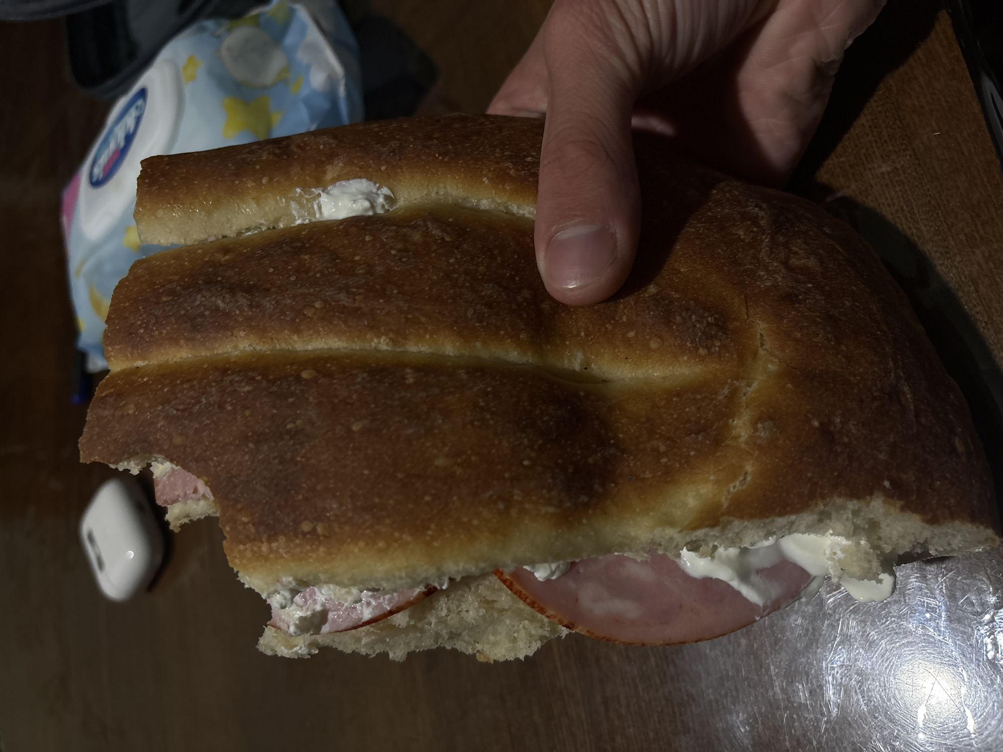 A partially eaten sandwich is held in someone's hand. The sandwich consists of two halves of brown bread with visible filling of ham and a white spread (possibly cream cheese). A bite has been taken out of the left side of the sandwich. In the background, there are baby wipes and the surface appears to be a wooden table. There is a pair of wireless earbuds to the left of the sandwich. The location of Yerevan, Armenia is not discernible from the image.