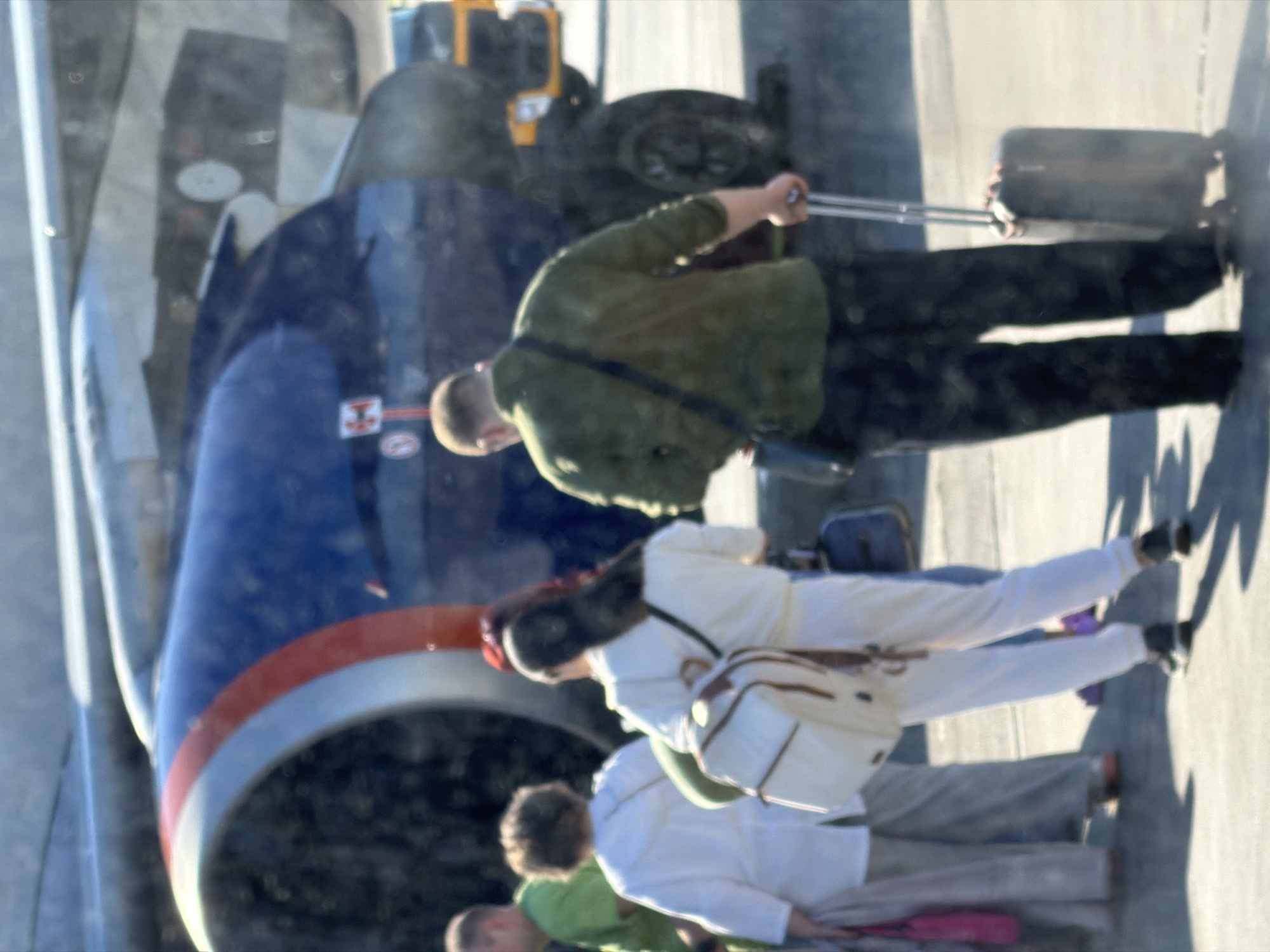 The image depicts an outdoor scene at an airport, likely within the city of Khimki, Russia, given its proximity to Sheremetyevo International Airport. In the foreground, four individuals are visible from the waist down, appearing to be passengers preparing to board an aircraft. The most prominent figure on the right is a person wearing dark pants and a green camouflage-patterned jacket, pulling a dark rolling suitcase with a silver handle. To their left, a person in white pants and a white top carries a cream-colored backpack with brown trim, slung over their shoulder. Behind them, another individual in a white top and grey pants is partially visible. Further to the left, a person in a green shirt is seen from the side. Dominating the background is the engine cowling of a large aircraft, characterized by its blue exterior with red and white stripes. The environment appears to be an airport tarmac, with the ground surface showing visible texture. The lighting suggests it is daytime, with shadows cast on the ground. The overall impression is one of passengers navigating an airport environment.