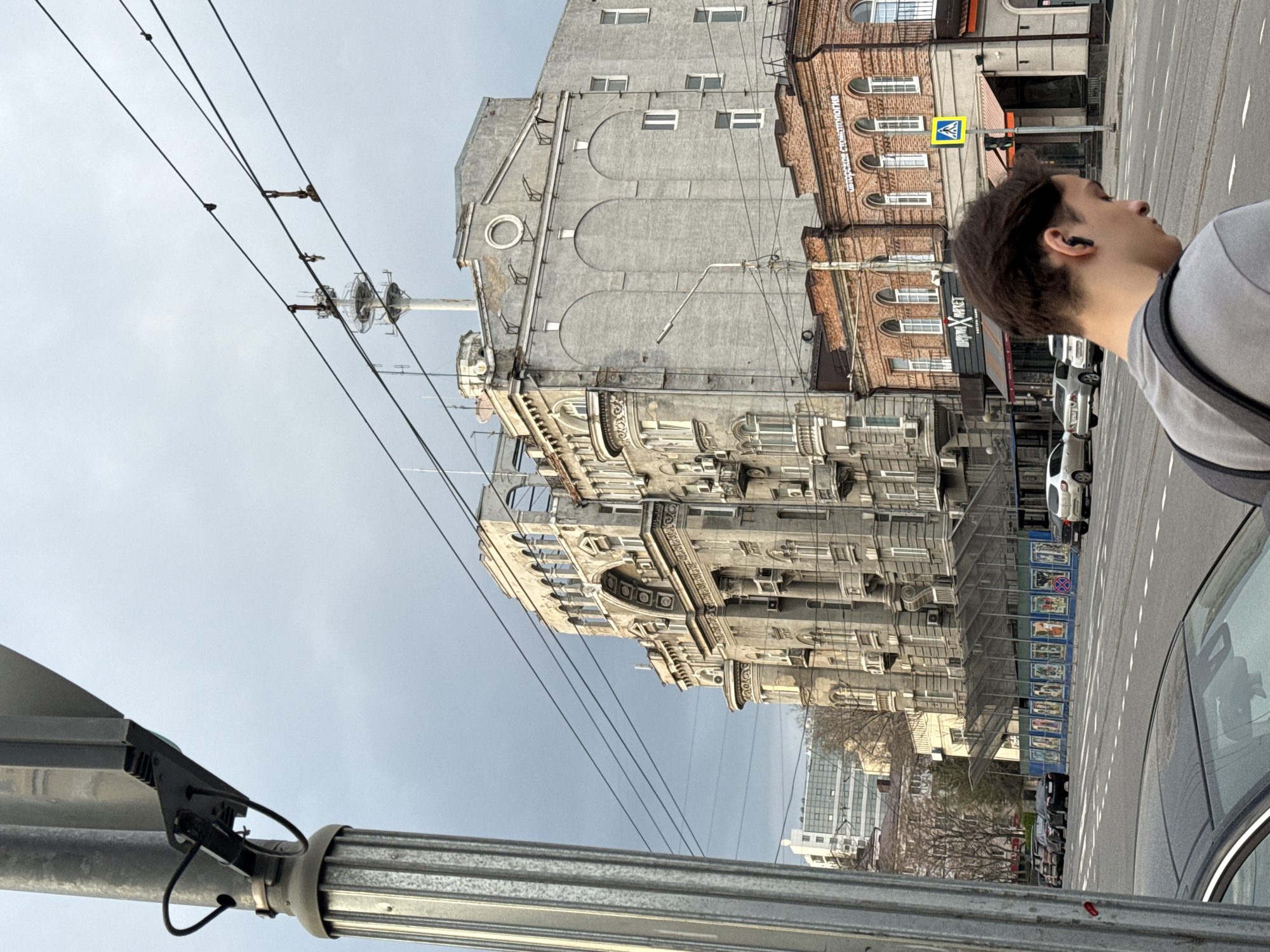The image shows a street scene in Rostov-on-Don, Russia, captured from a low angle. A male person is partially visible in the right side of the frame. He has short dark hair and is wearing a grey top with a dark strap. He is looking ahead. Adjacent to him, cars are parked along the roadside. The street is clearly marked with white lane dividers. A multi-story building stands prominently, showcasing a blend of grey and brick architectural styles. The building's facade is adorned with decorative elements, and windows are visible across its height. Construction work is taking place next to the building. Power lines are crisscrossing the sky. The sky is overcast, suggesting a cloudy day. A metallic pole with attached equipment is visible in the foreground, adding depth to the scene.