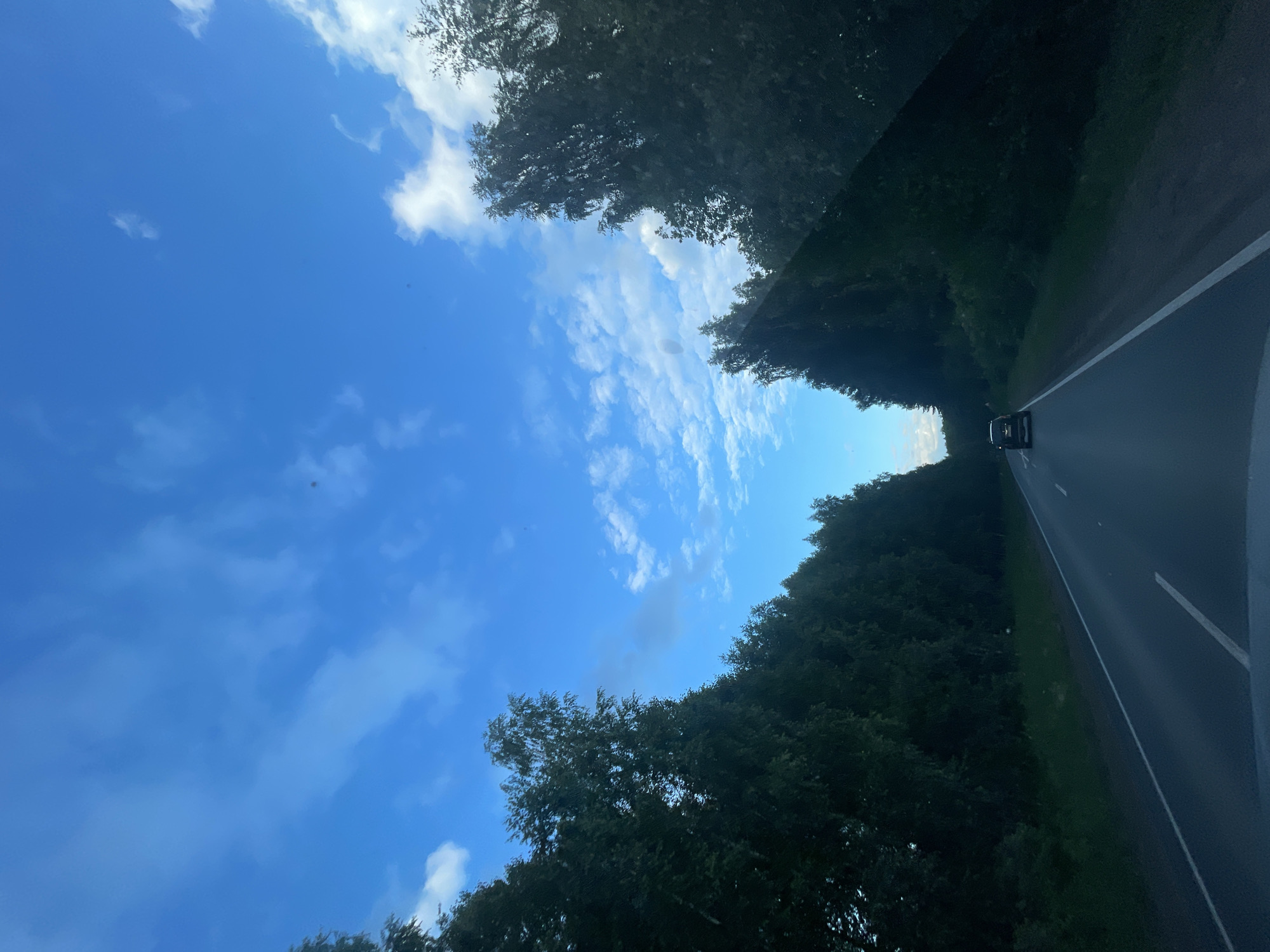 This image depicts a road curving through a dense forest under a bright blue sky with scattered white clouds. The perspective is from inside a vehicle, looking forward and slightly upwards. A single black car is visible in the distance on the road. The trees lining the road are lush and green, suggesting a warm season. The lighting indicates it is daytime, likely late morning or afternoon, with the sun creating a slight glare on the vehicle's windshield which is visible in the foreground. The scene conveys a sense of travel and a serene, natural environment. There is no visible text in the image.