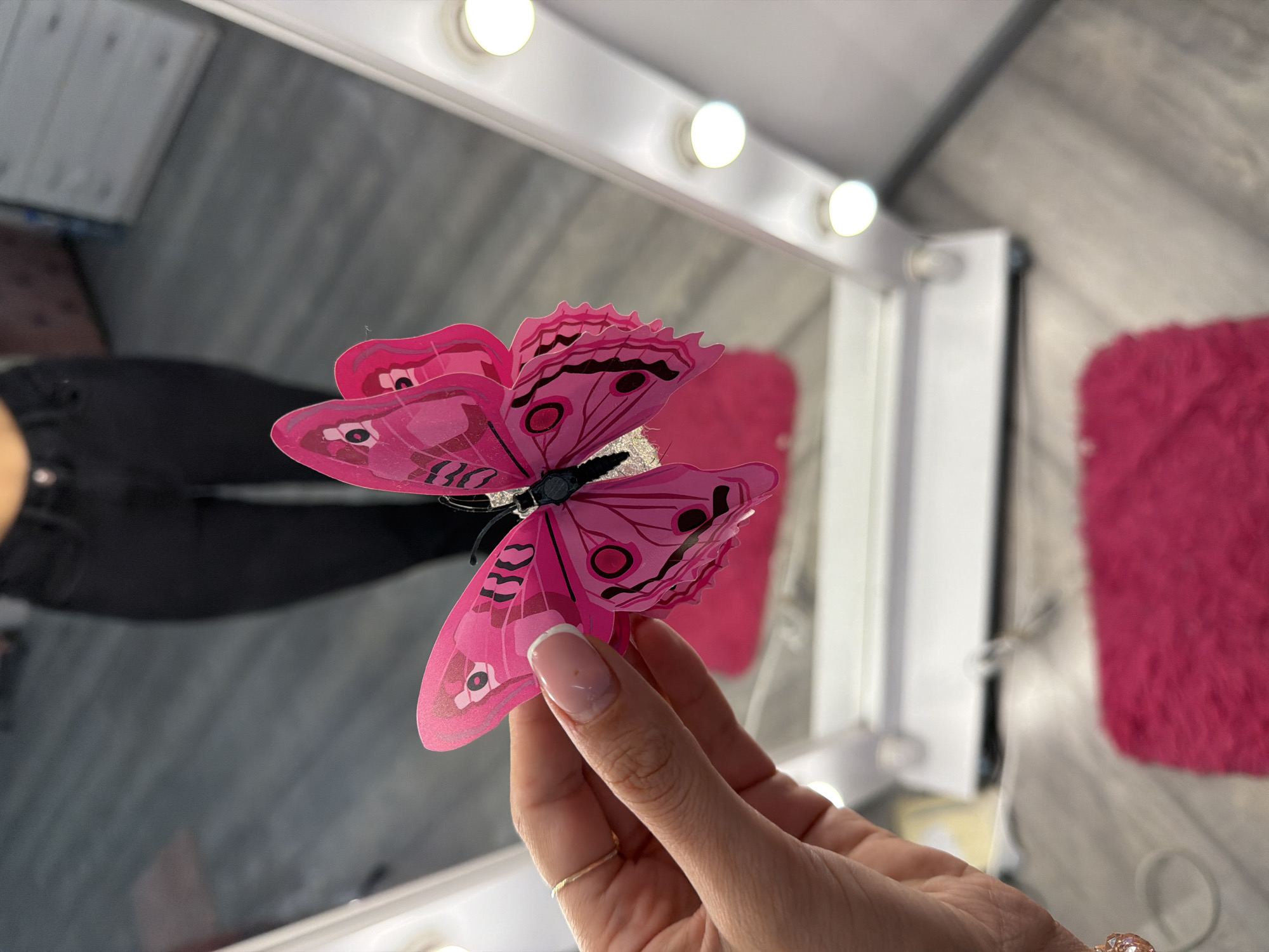 A close-up shot focuses on a hand holding a bright pink, decorative butterfly. The butterfly is positioned in the foreground, with its wings spread. The hand is partially visible, with a gold ring on one finger and neatly manicured nails. In the background, a person wearing black pants is reflected in a large mirror. The mirror appears to be part of a vanity or dressing area, as evidenced by the row of illuminated bulbs lining its frame. The floor has a light gray wood-grain pattern, with a vibrant pink, shag rug visible in the lower portion of the image. The overall setting suggests an indoor, personal space, possibly a dressing room or bedroom. There is no indication of the specific location within the city of Kyiv, Ukraine.