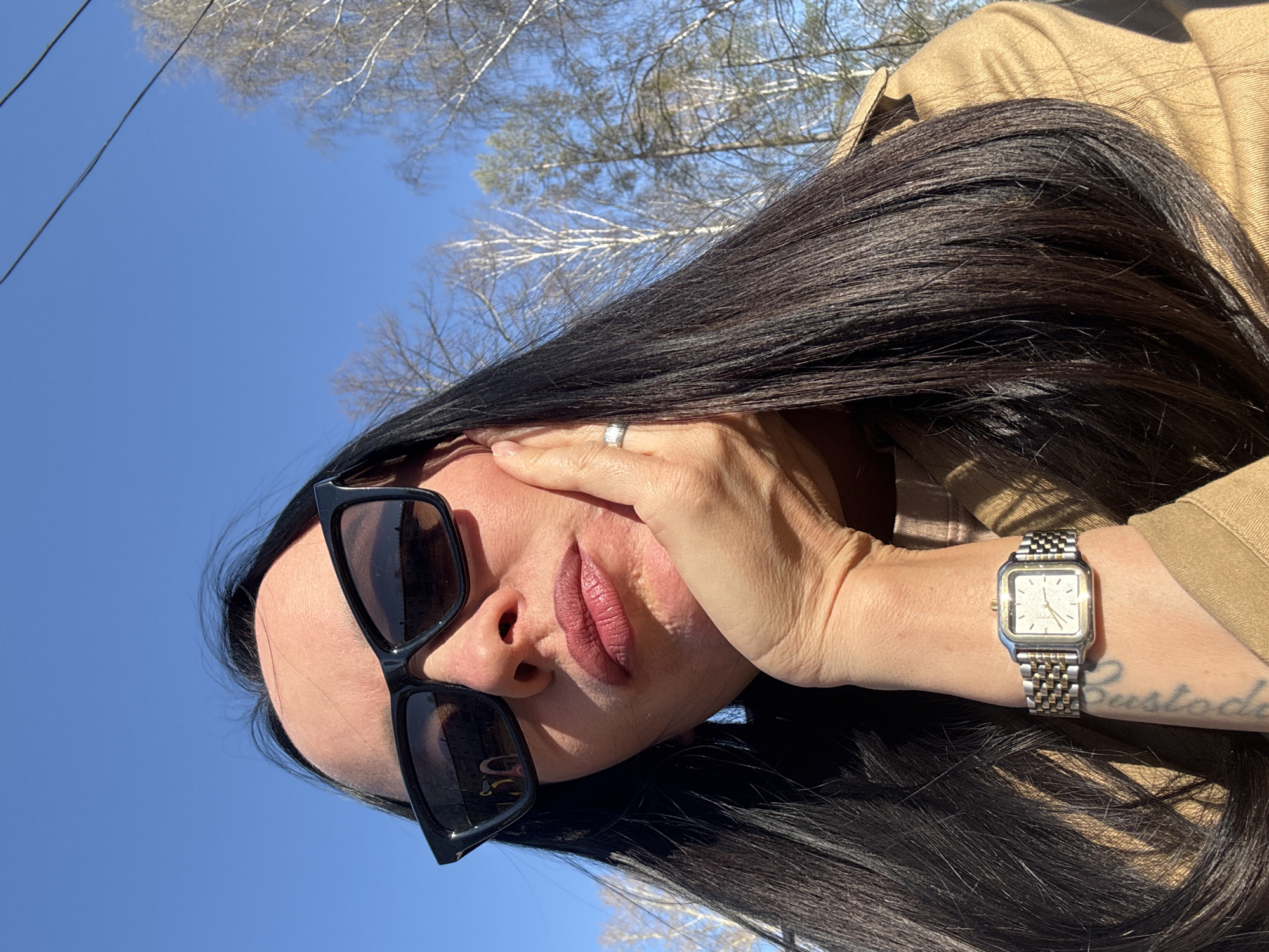 A woman is seen from a low angle, with a backdrop of a clear blue sky and bare tree branches. She is wearing dark sunglasses and a tan-colored jacket. Her dark hair is styled and flowing. She is wearing a silver watch on her left wrist. Her right hand is positioned near her face, and a ring is visible on her finger. The tattoo can be seen on the left wrist. The location cannot be identified as Chaikovsky, Russia.