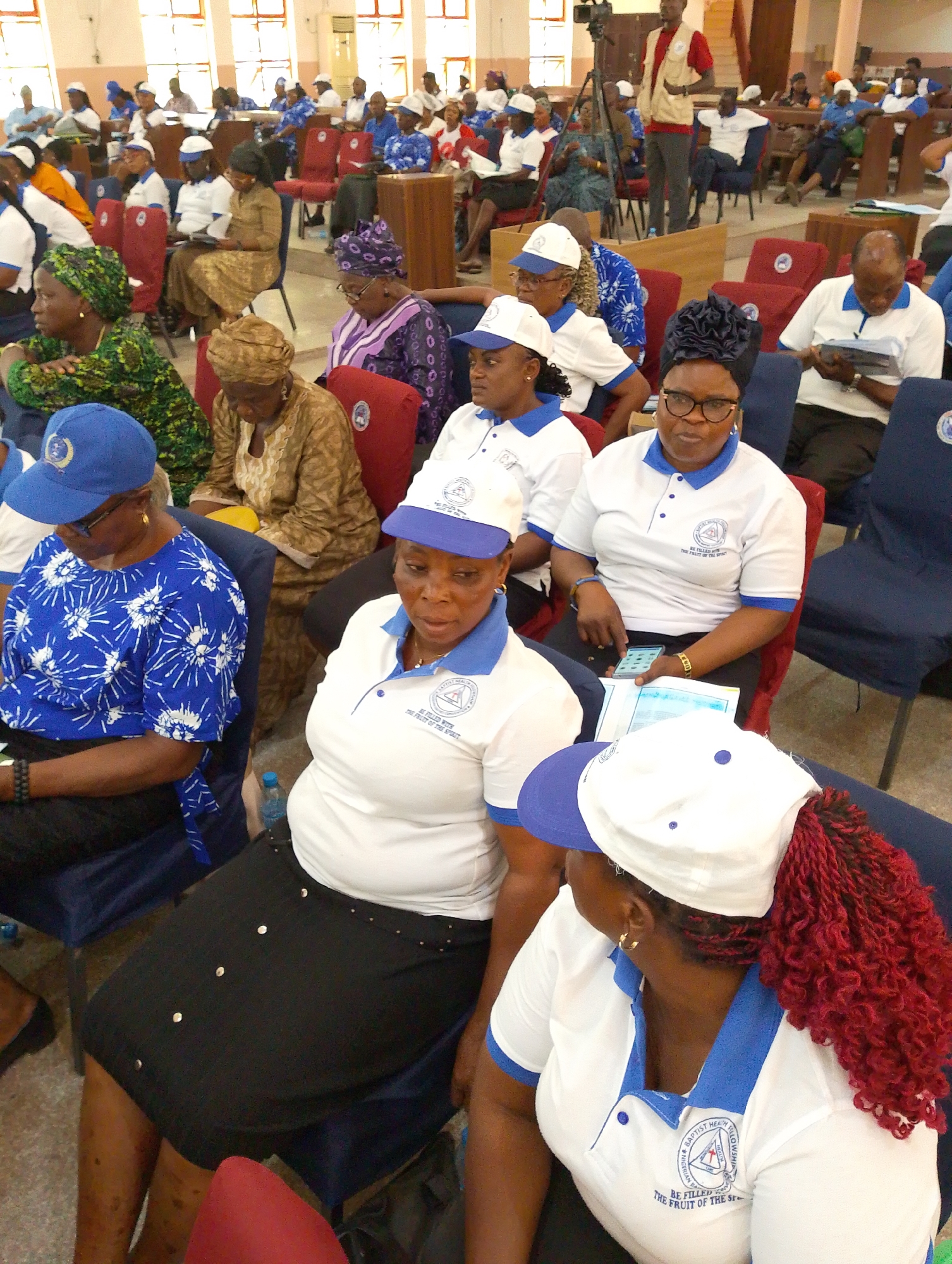 This image captures an indoor gathering or conference, likely in a hall or auditorium, in Kubwa, Nigeria. Numerous people, predominantly women, are seated in rows of blue and red chairs.
Many attendees wear a uniform consisting of white polo shirts with blue collars and trim, paired with white baseball caps featuring blue visors. The logo visible on these shirts and caps reads "NIGERIAN BAPTIST HEALTH FELLOWSHIP" and includes the motto "BE FILLED WITH THE FRUIT OF THE SPIRIT." Several women also wear traditional Nigerian head wraps in various colors, such as purple, green, and brown, with patterned fabrics.
In the foreground, a woman on the right wears the uniform and has distinctive long, red braided hair gathered under her cap. Another woman in the foreground is dressed in a blue and white patterned top with a matching blue cap. A woman in the center, also in uniform, looks towards the camera while holding a mobile phone and documents. Throughout the room, people are engaged in the event, some listening attentively, others looking at documents, and a few interacting with their phones. A man in a red vest is visible standing further back near a camera setup, suggesting the event is being recorded or managed. The setting is brightly lit, with tall windows visible in the background. The overall mood appears to be one of calm attention and participation.