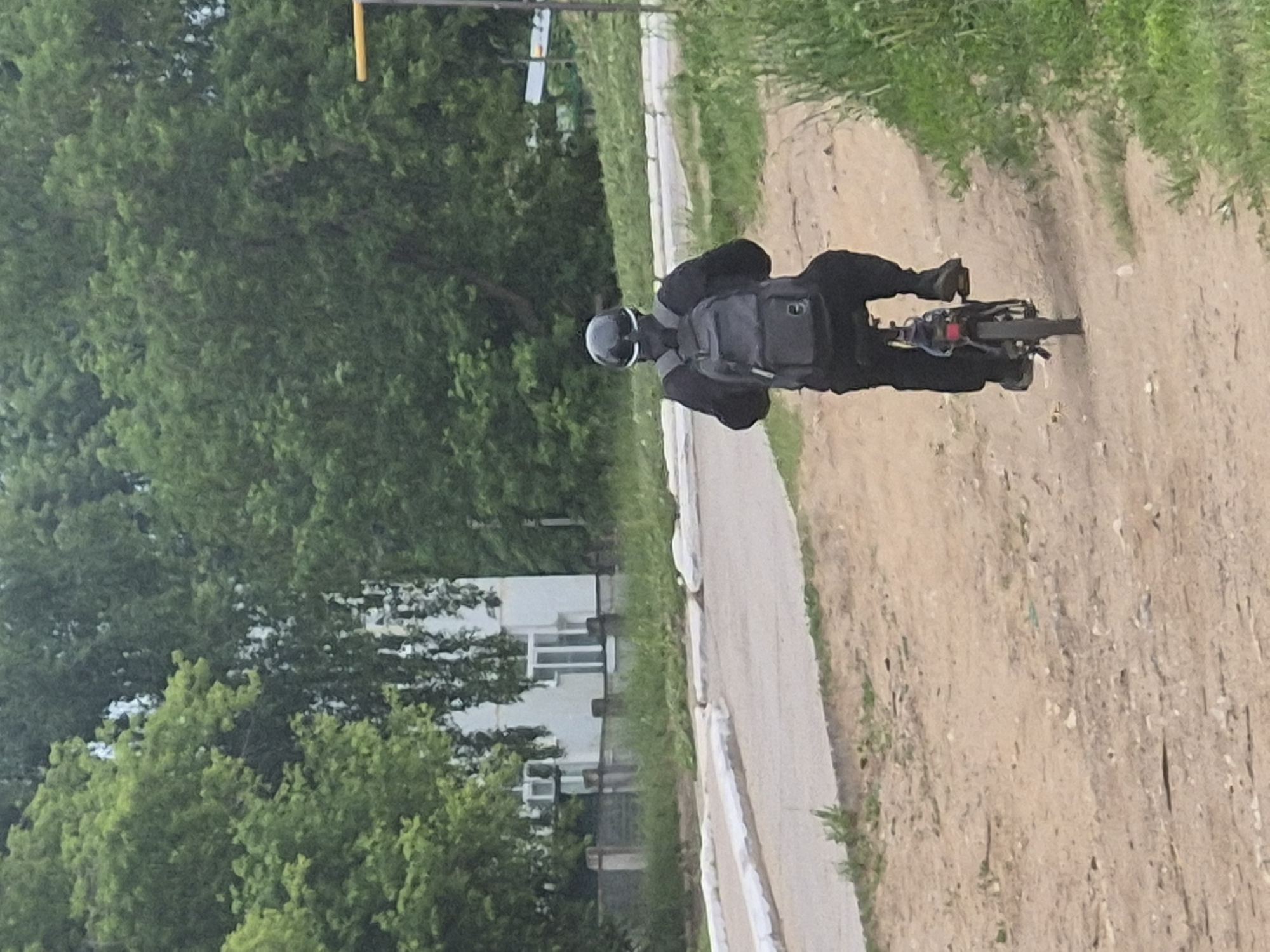 An overhead view captures a person riding an electric scooter or small motorcycle on a dirt path in Chaikovsky, Russia, during daytime. The rider is dressed in black clothing, including a jacket and trousers, paired with a black backpack and a light-colored helmet. They are positioned on the dark-colored vehicle, actively steering.
To the left of the dirt path, a paved sidewalk or narrower asphalt path runs parallel, bordered by a distinct white curb. Lush green trees and vegetation form a dense backdrop, indicating a natural or suburban setting. Through the trees in the mid-ground, a white building with multiple windows is visible, suggesting a residential or public structure. A yellow pole, possibly part of a sign or utility structure, is seen in the upper right portion of the frame, and a faint, unidentifiable sign is visible further back among the trees. The weather appears clear with good visibility. No discernible text is visible in the image.