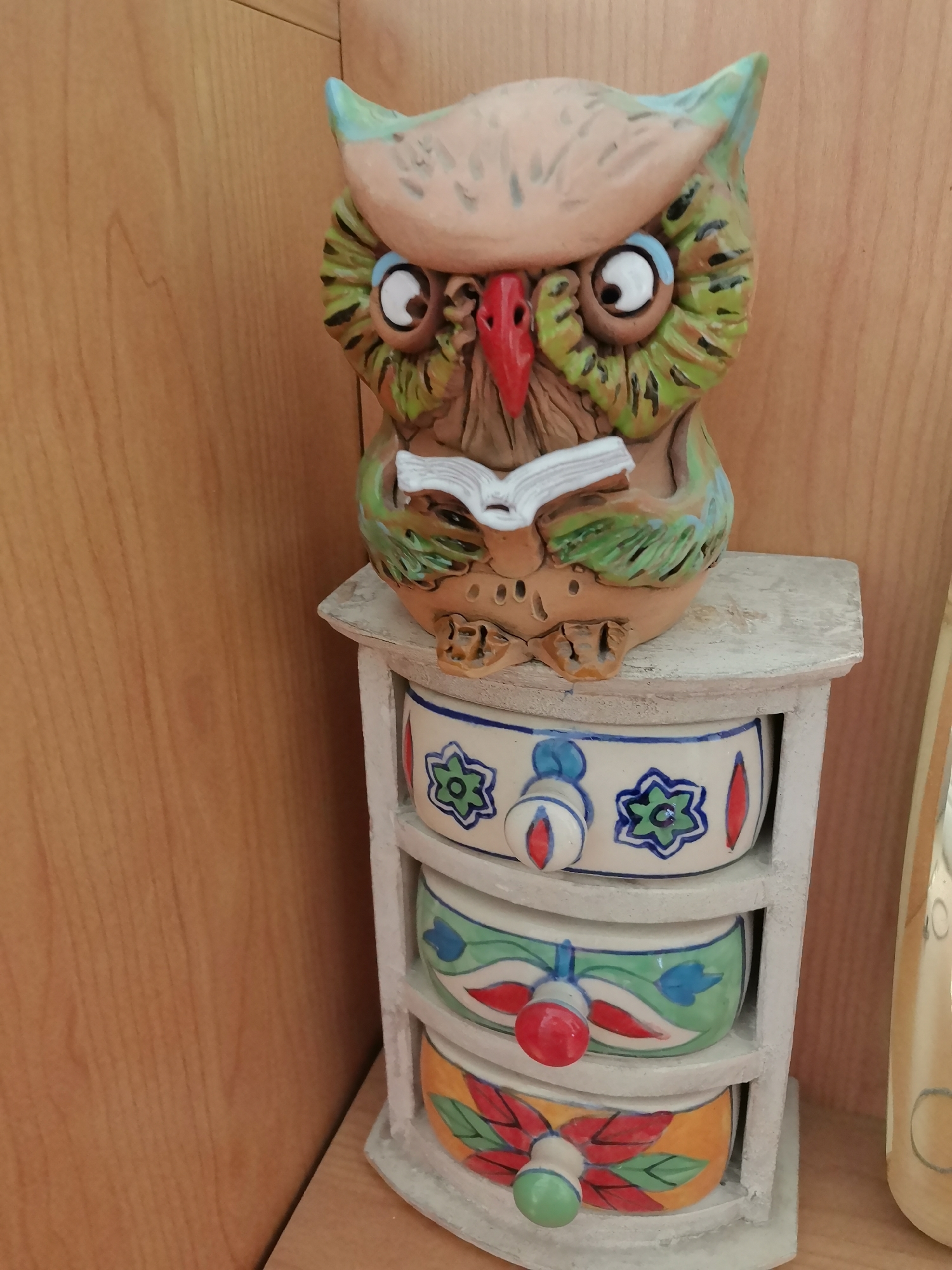 An artistic ceramic owl figurine, depicted as reading a book, rests atop a small, white, three-drawer cabinet. The owl is colorful, with green and brown feathers, large white eyes with blue accents, and a red beak. Each drawer of the cabinet is decorated with unique, hand-painted floral patterns in various colors, including blue, green, red, and yellow. The cabinet and owl are placed on a light-colored wooden surface. To the right of the cabinet, a portion of a tall, light-colored ceramic vase with circular patterns is visible. The background consists of a warm-toned wooden wall. There are no people or any indication of the city of Oryol, Russia, present in this image.