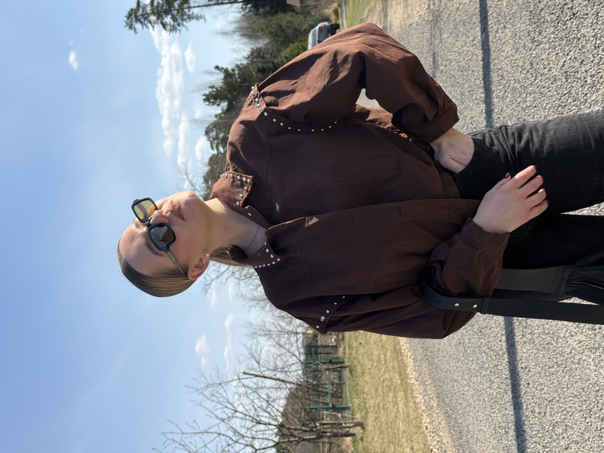 A woman wearing a brown jacket with silver stud detailing and black jeans stands on a gray asphalt road. She has on sunglasses and is looking upwards. A black belt is visible around her waist. The background features bare trees, patches of grass, and a blue sky. The location is likely outdoors, possibly in or near the city of Dreiliņi, Latvia, based on the broader context of the request.
