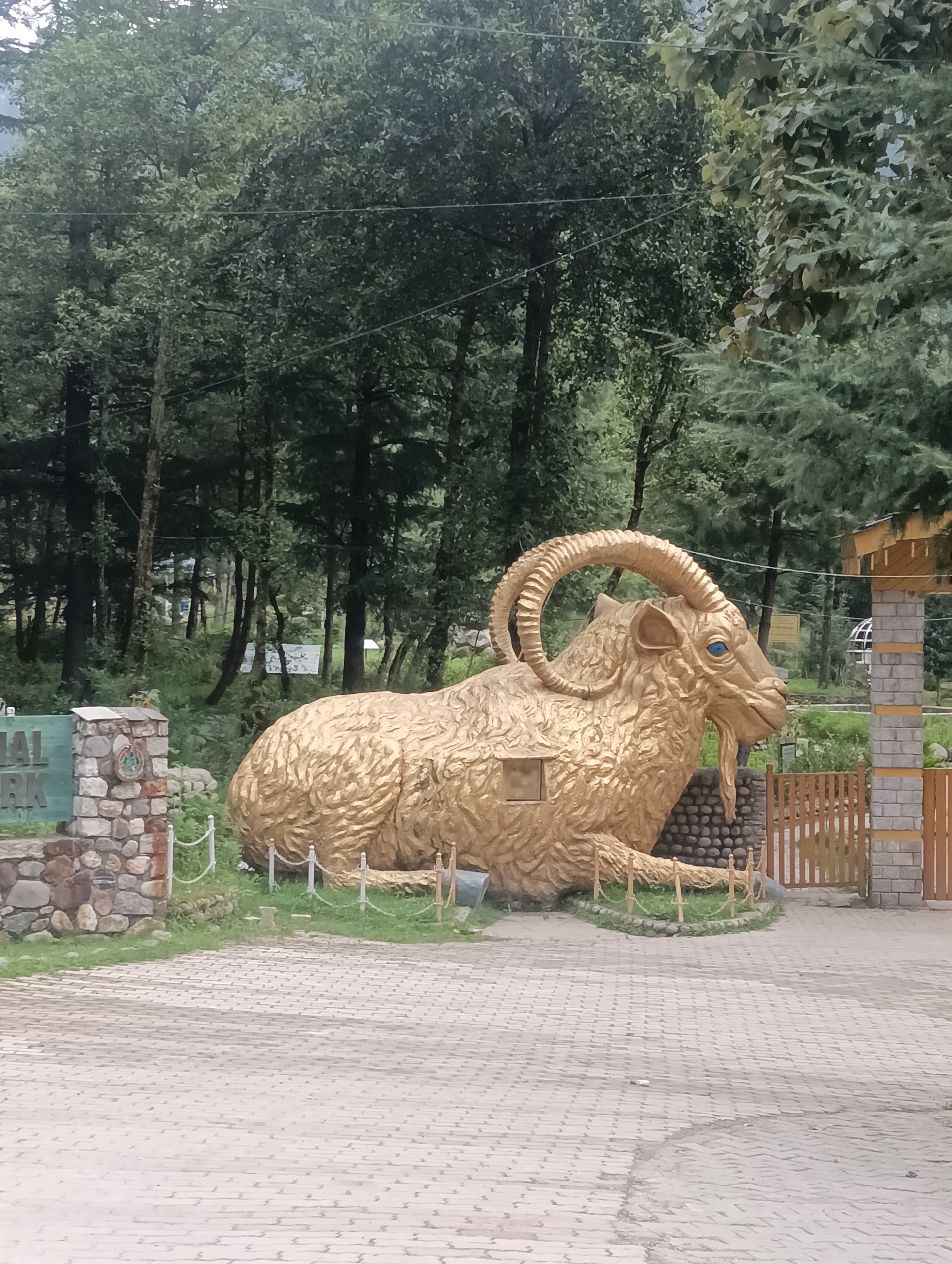 This is an outdoor, full-shot image of a large, golden statue of a ibex, a type of wild goat, located in Manali, India. The statue is positioned in the center of the frame, facing to the right. It has large, curved horns and striking blue eyes. The statue is textured to resemble fur. In front of the statue, there is a paved area made of light-colored rectangular bricks. To the left of the statue, there is a stone monument with a sign that reads "NATIONAL PARK." The monument is made of rough-hewn stones and has a circular emblem on it. To the right of the statue, there is a wooden gate and a brick structure that appears to be an entrance. The background consists of a dense forest with tall, green trees. The overall scene suggests a tourist attraction or an entrance to a park. The lighting is overcast, contributing to the muted colors of the foliage and the environment.