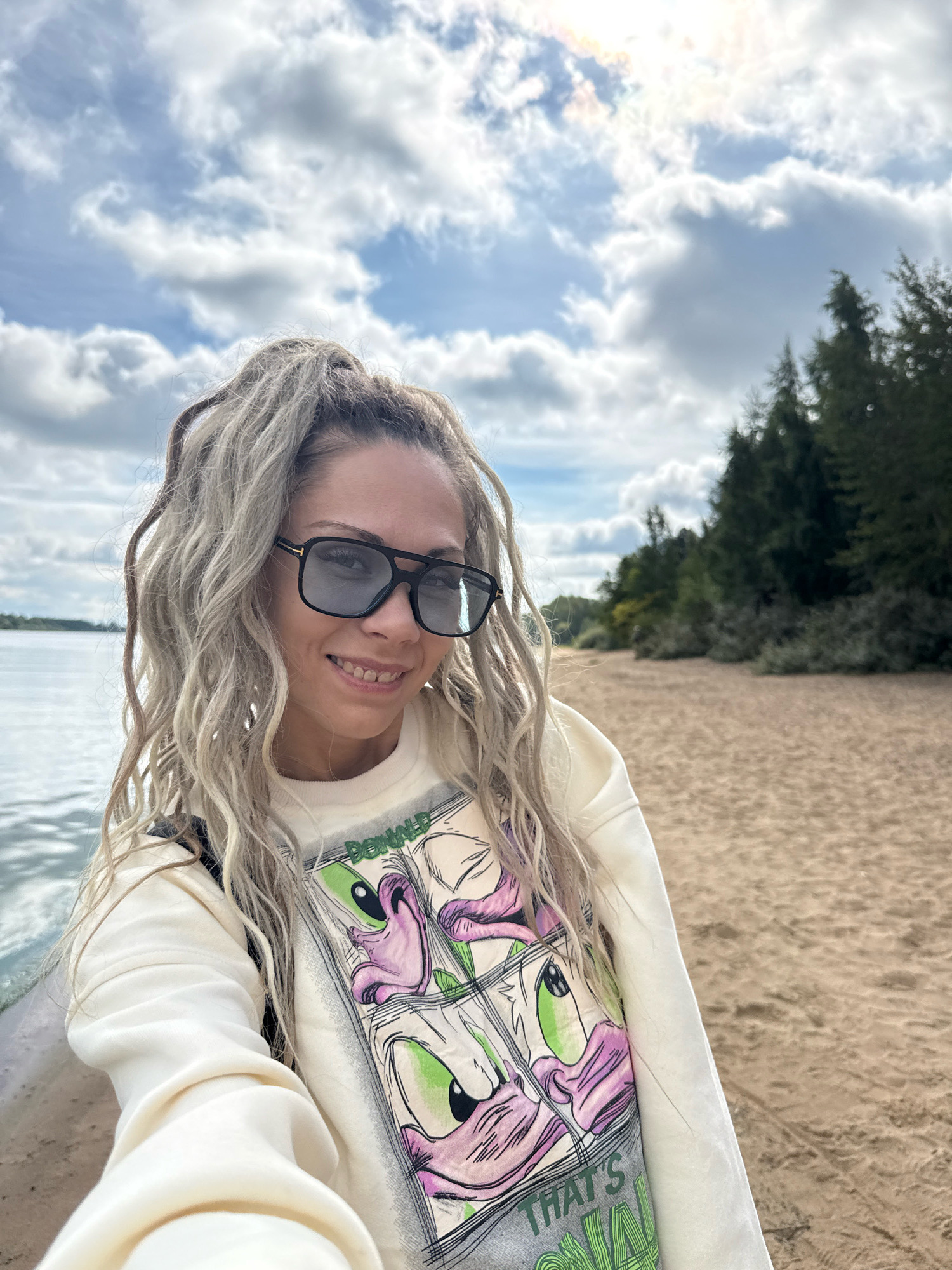 A selfie taken by a woman with blonde dreadlocks and sunglasses, smiling at the camera. She is wearing a cream-colored sweatshirt with a graphic print of cartoon characters. The background shows a sandy beach, a body of water, and trees under a cloudy sky. The location is identified as Rusanovo, Russia.