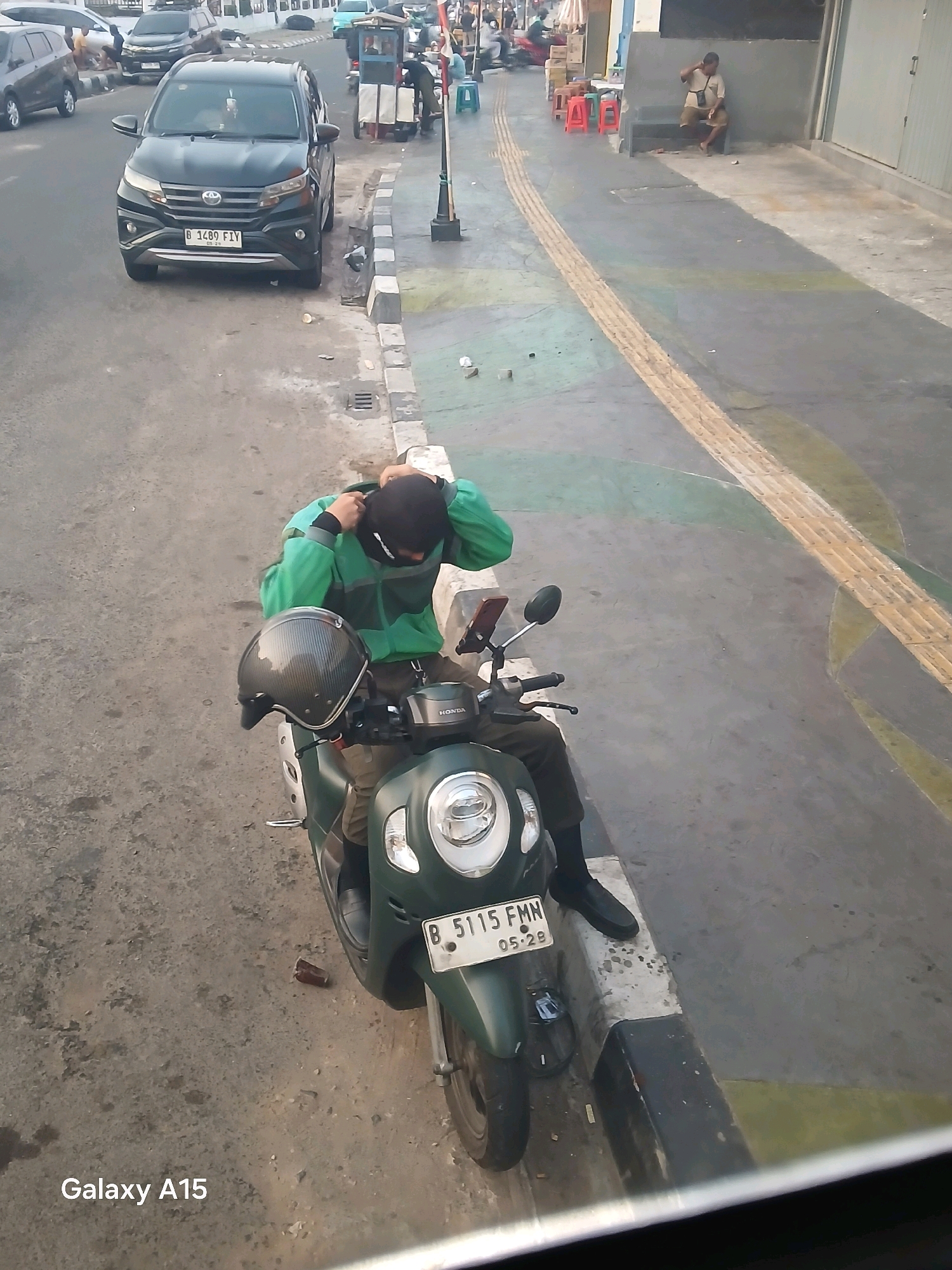 A person is sitting on a green scooter on the side of a street in Bekasi, Indonesia. The person is wearing a green jacket, dark pants, and a helmet. They are adjusting the helmet strap. The scooter has a license plate that reads "B 5115 FMN". In the background, there is a black car with a license plate that reads "B 1489 FIV", other vehicles, a sidewalk with yellow tactile paving, and some buildings. A man is sitting on the stairs of one of the buildings. The weather appears to be sunny.