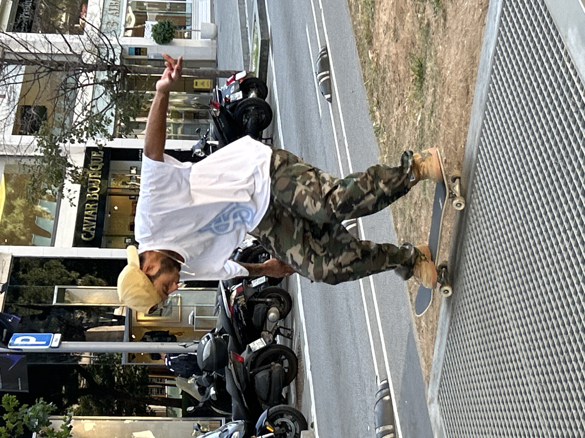 An urban street scene in Barcelona, Spain, captured during daytime under clear weather. The main subject is a person, likely a male, engaged in skateboarding. He is positioned on a skateboard with his left foot on the board and his right foot slightly off, appearing to balance or perform a trick. He wears a white t-shirt, camouflage pattern pants, tan shoes, and a light-colored baseball cap worn backward. His right arm is raised, with his thumb and pinky finger extended in a "shaka" or similar gesture.
The setting includes a multi-lane asphalt road with white markings and several parked motorcycles or scooters to the left of the skater. Adjacent to the road are multi-story buildings with large glass windows, suggesting commercial establishments. Trees with sparse foliage are visible between the buildings and the road. A metal grate or grid structure runs alongside what appears to be a raised sidewalk or platform on the right side of the image, next to a patch of dry dirt and grass.
Visible text includes "CAVIAR BOUTIQUE" and partially "VIAR BOUTIQUE" on a storefront, "3 ALBA" on another building, and a blue "P" parking sign. The image captures a dynamic moment in a busy urban environment.