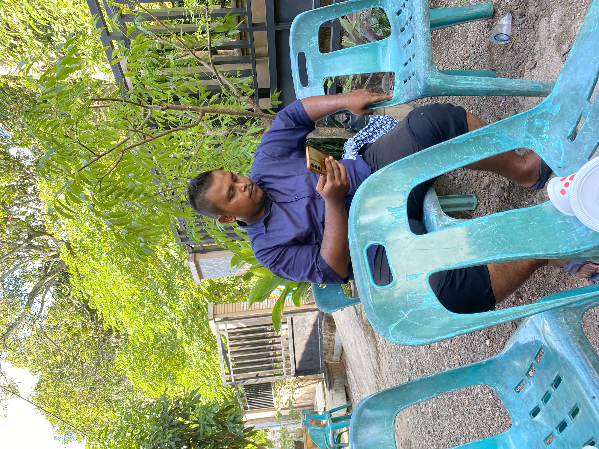 In an outdoor setting, a man with a mohawk hairstyle is seated on a teal plastic chair, holding a gold smartphone in his right hand. He is wearing a dark blue collared shirt and black shorts. The man is looking down at the phone, and his left hand rests on the armrest of the chair. In front of him, another teal plastic chair is visible. A blue and white patterned cloth is draped over the back of the chair the man is sitting on. The ground is covered with gravel, and there are several trees and a black metal fence in the background. The scene appears to be in a residential area with some sunlight filtering through the trees.