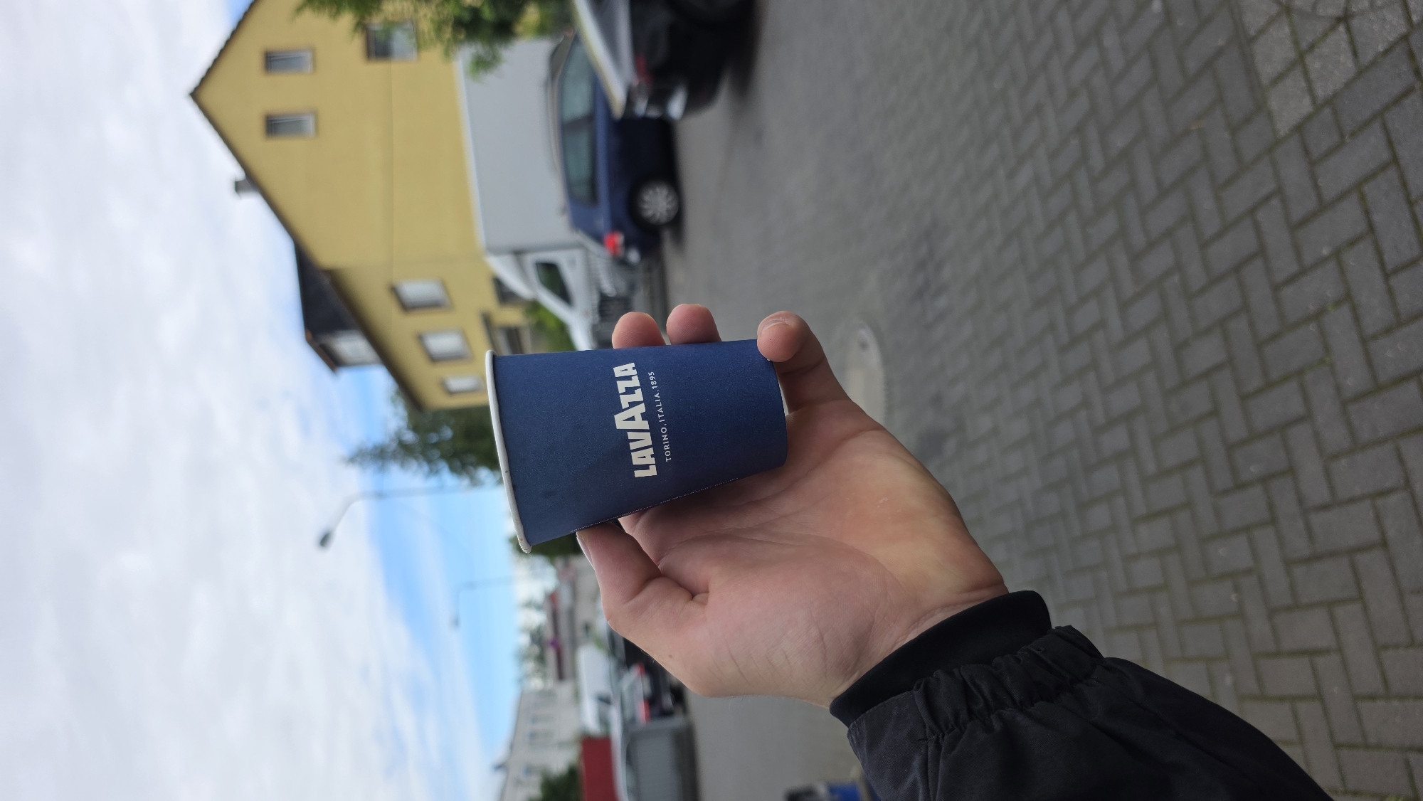 A person's hand is holding a blue paper coffee cup with the Lavazza logo and "TORINO, ITALIA, 1895" printed in white. The cup is positioned in the foreground, with the person's arm and jacket sleeve visible on the left side of the frame. In the background, there is a yellow building with windows, a white van, a dark blue car, and a black car parked on a paved surface. The sky is overcast with clouds. The location is identified as an outdoor urban setting.