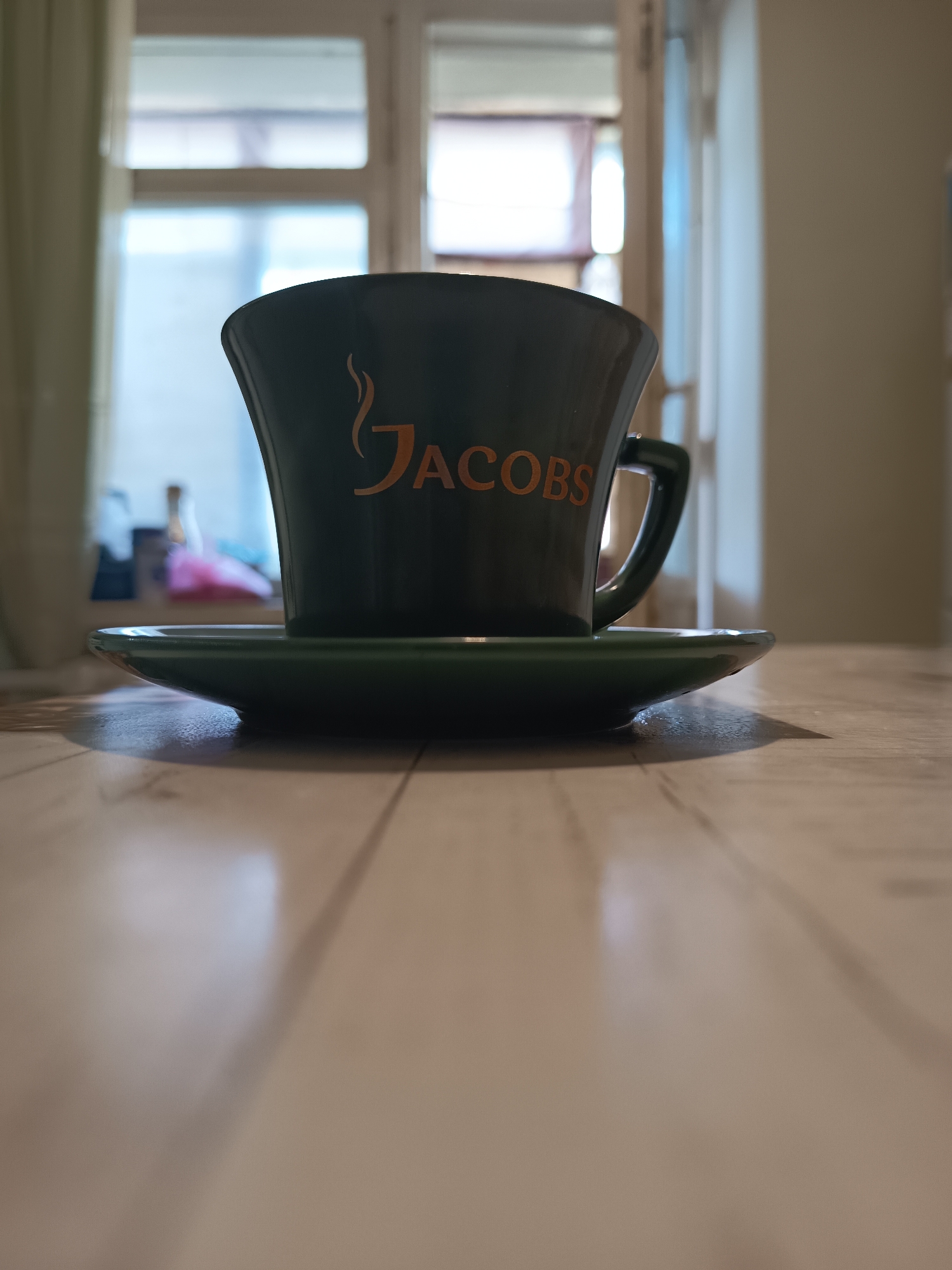 A low-angle, close-up shot shows a dark green coffee mug with the orange Jacobs logo on it, sitting on a matching saucer. The mug and saucer are placed on a light-colored wooden table. In the blurred background, a window with vertical blinds is visible, suggesting an indoor setting, possibly a cafe or home. The lighting creates a shadow from the mug and saucer onto the table. There are no people or animals visible in the image. The specific location in Izmail, Ukraine, cannot be determined from the visual information provided.
