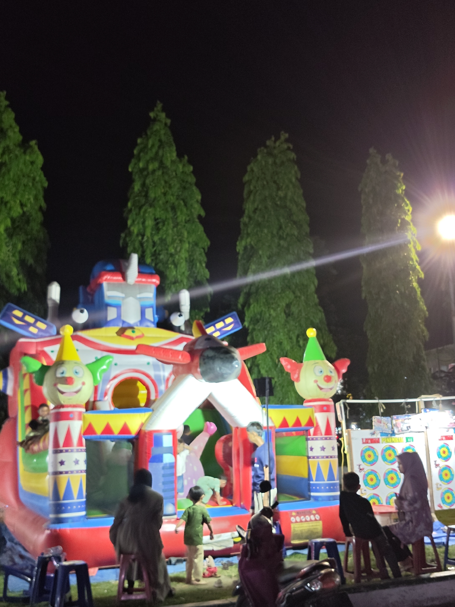 The image shows an inflatable playground at night in Lhokseumawe, Indonesia. The playground is brightly colored with clown faces and geometric shapes. Children and adults are near the structure, some interacting with it. There are tall trees in the background and a light source illuminating the scene. People sit on stools near a game with targets. A motorcycle is parked in the foreground.