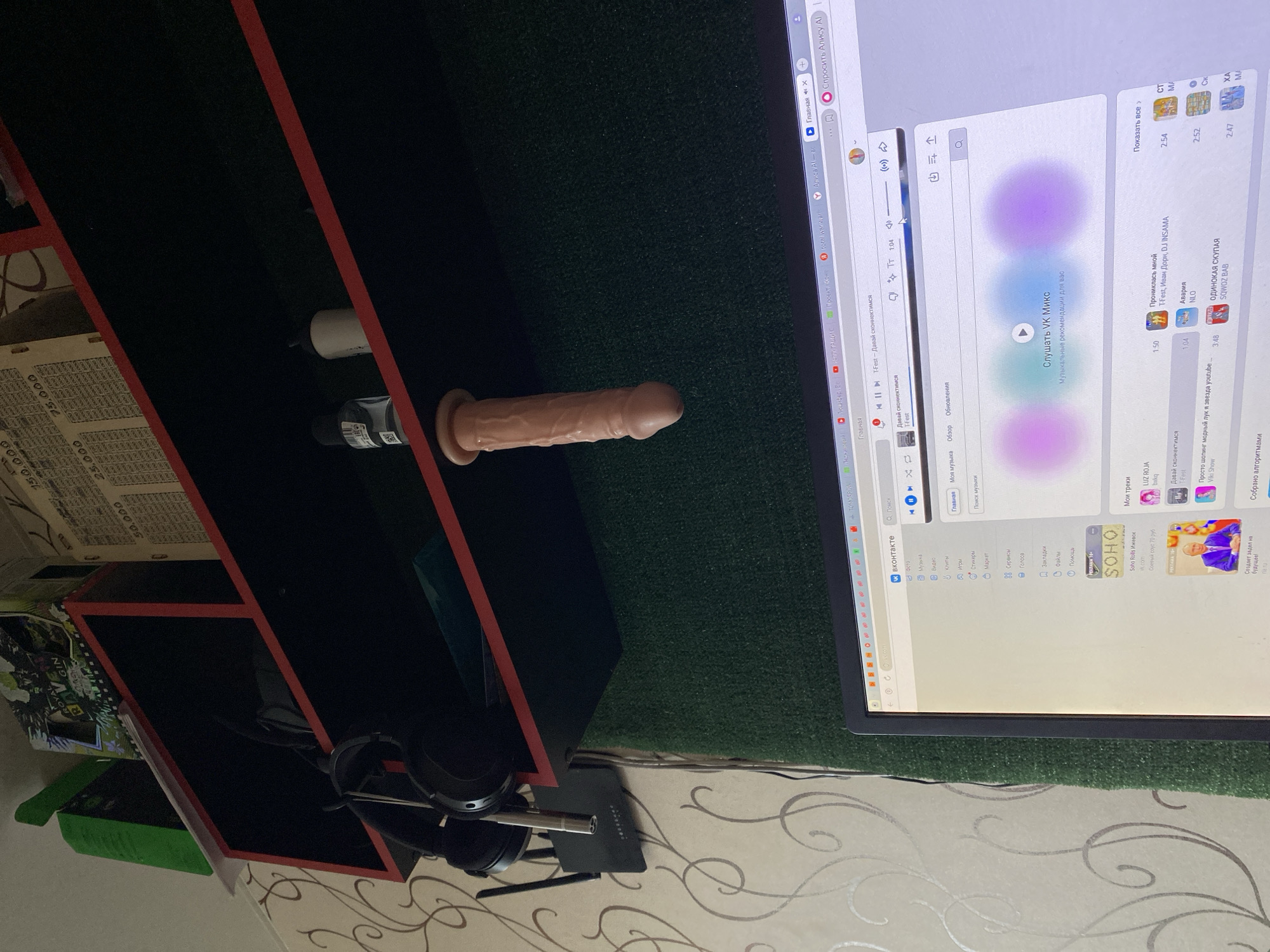 A room has a computer and a black shelf with a red trim. The shelf has some miscellaneous objects including headphones, papers, and personal items. A rubber dildo is placed on the shelf near the computer screen. The computer screen shows the Russian VK music service. The location is not discernible to be Izhevsk, Russia.
