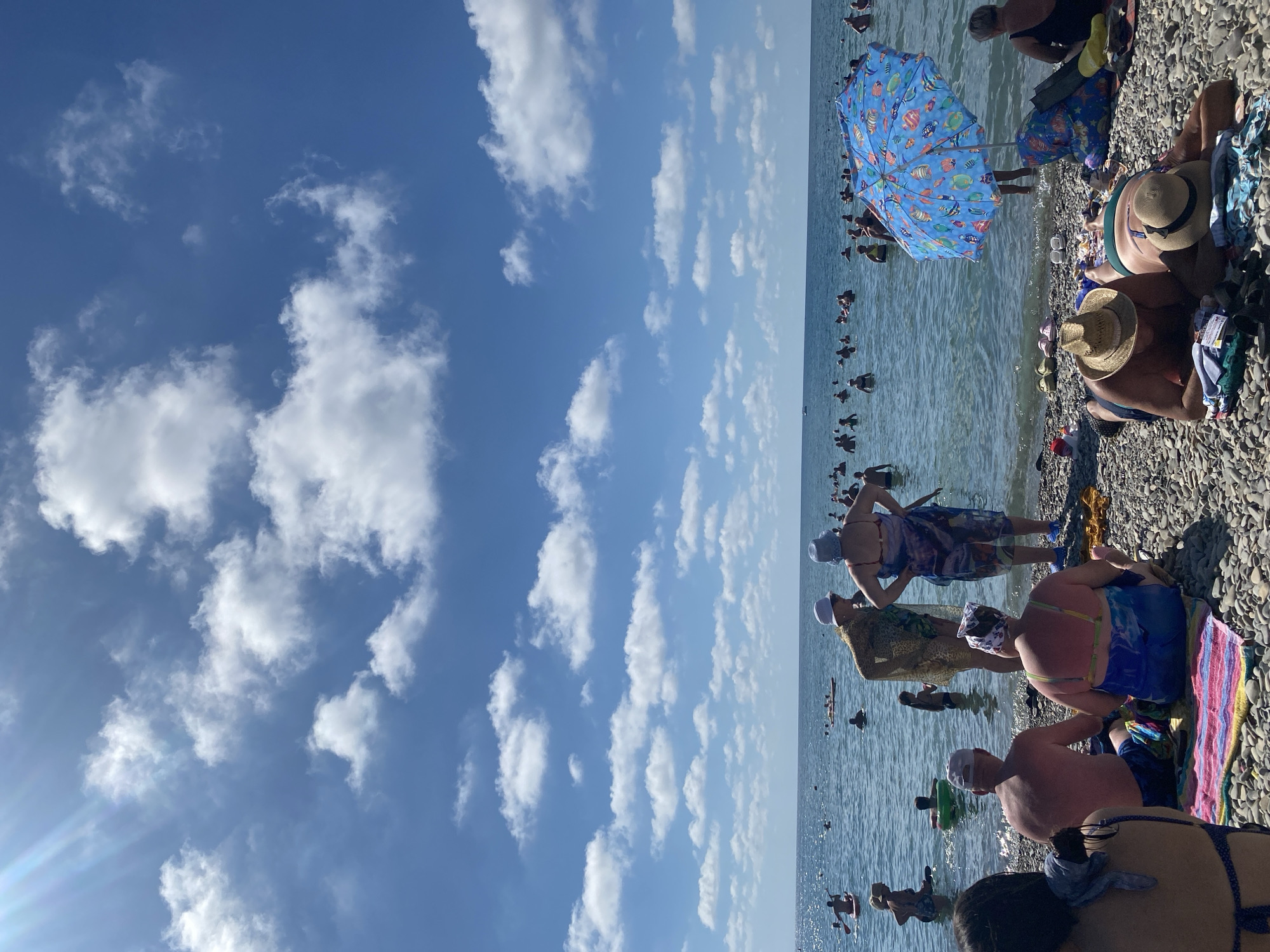 This is a vertical, eye-level, outdoor shot taken on a sunny day at a pebble beach in Arkhipo-Osipovka, Russia. The sky is a vibrant blue, filled with scattered white cumulus clouds, and a sunbeam extends from the upper left corner. The sea is calm, with a slight shimmer on the surface, and populated by numerous people swimming and wading.
In the foreground, several individuals are seen on the beach. Two people are lying down on towels, facing away from the camera. One is wearing a straw hat and the other a light-colored hat. Another couple is seated on a towel, also facing away, with one person wearing a white cap and the other a patterned bandana. To the right, a bright blue umbrella with a fish pattern is open, and a woman with short grey hair is sitting nearby.
Further back, a group of people are standing in the shallow water. Two individuals in the center are conversing, one wearing a patterned wrap and a white hat, the other a blue wrap and a blue hat. More people are visible in the water further out, some on inflatable floats. A person on a paddleboard is visible in the left midground. The beach itself is covered in smooth, grey pebbles.