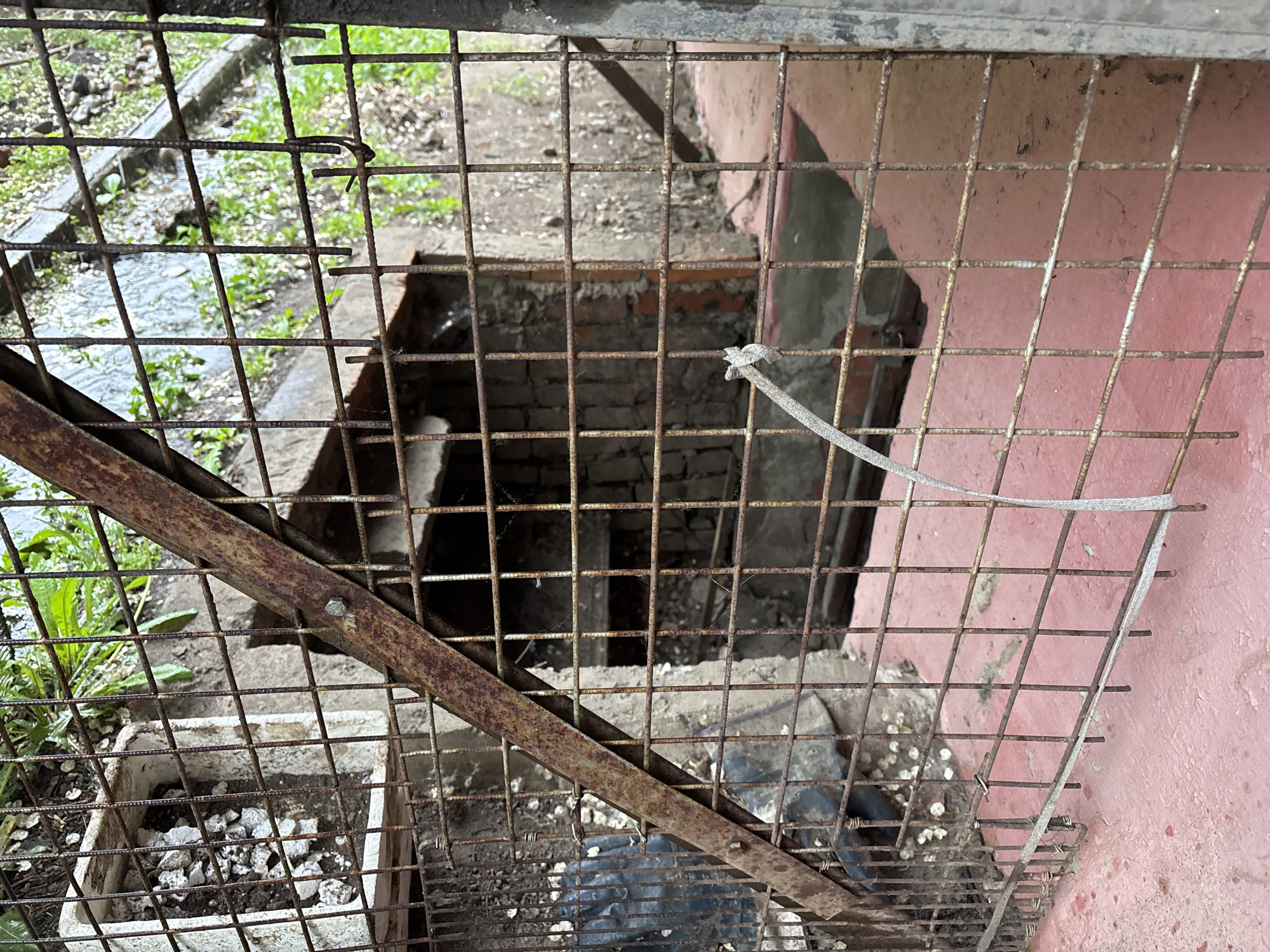 A ground-level view through a rusty, wire mesh fence. Beyond the fence is a brick-lined opening leading to an indeterminate underground space. A pink painted wall is visible to the right of the opening. The fence is secured by a loosely tied white strap. A container filled with rocks and soil is visible behind the fence. The location appears to be an outdoor, urban environment in Unknown, Unknown.