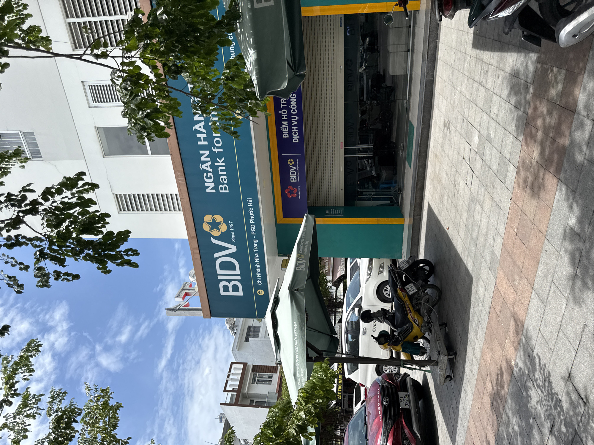 The image shows the exterior of a BIDV bank branch in Nha Trang, Vietnam, on a sunny day. The bank has a teal facade with prominent BIDV branding. The entrance is a large glass door with a rolling security shutter pulled down. Above the entrance, there is a sign that reads "ĐIỂM HỖ TRỢ DỊCH VỤ CÔNG" (Public Service Support Point).
In front of the bank, a large green umbrella provides shade. Parked on the sidewalk are several vehicles, including a red SUV with the license plate "79A 328.64" and two motorcycles. The sidewalk is paved with gray and reddish tiles. Trees with green leaves hang over the scene, casting dappled shadows. In the background, there are other buildings, suggesting an urban environment. The sky is blue with scattered white clouds.