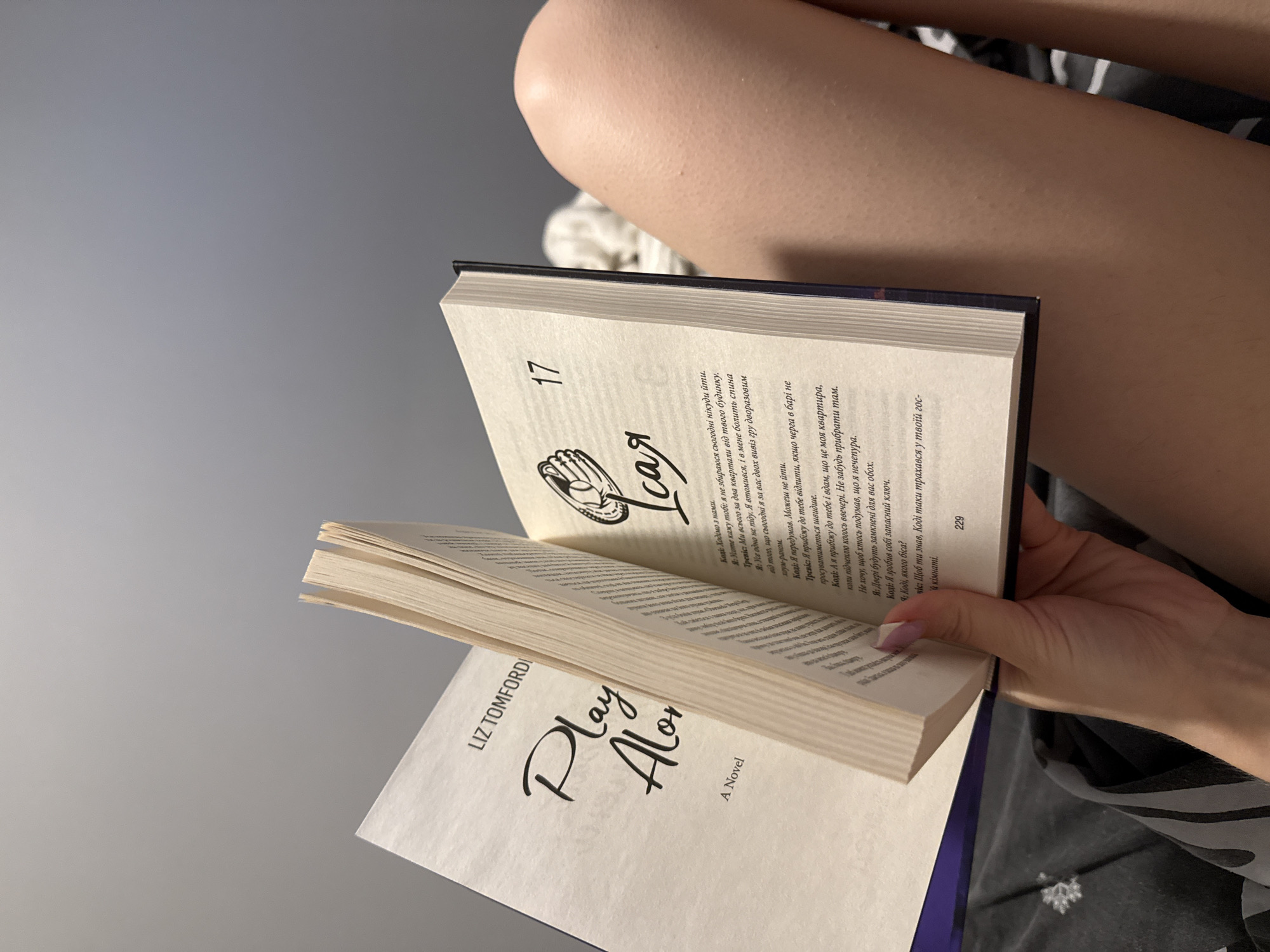 An open book titled "Play Alone" by Liz Tomfordi is held by a person's left hand, with the right page displaying the number "17" and some text in Ukrainian. The book is lying on a dark-patterned fabric, likely a bed or blanket, and a person's legs are visible on the right side of the frame. The background is a plain grey wall. The scene is an indoor setting, likely a bedroom, and no specific landmarks or locations in Łódź, Poland are depicted or suggested.