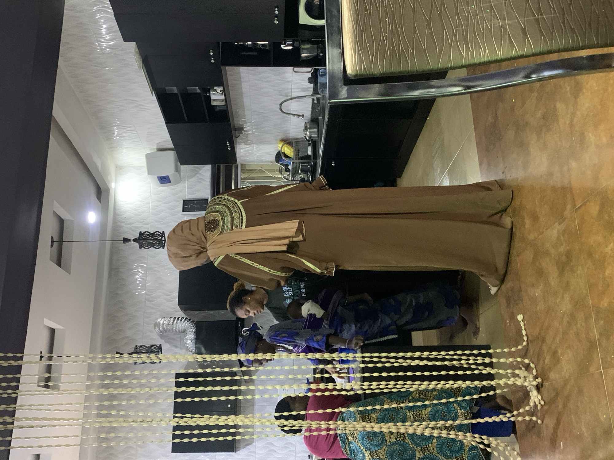 This image depicts an indoor domestic scene in Jos, Nigeria, featuring at least five individuals within a combined kitchen and common area.
In the immediate foreground, on the left, a decorative curtain composed of multiple vertical strands of off-white elongated beads hangs, partially obscuring the view. Some strands extend to the floor, where they are slightly tangled. The floor throughout the space is covered with large, square, brownish-orange ceramic tiles.
On the left side of the mid-ground, a person is partially visible behind the beaded curtain, wearing a blue and teal patterned lower garment and a pink upper garment. To their right, a woman in a blue patterned traditional dress with a matching headwrap stands barefoot, carrying an infant secured in a purple fabric wrap on her back. She is positioned facing a dark-colored kitchen counter. Adjacent to her, a woman with brown curly hair wears a dark green t-shirt with partially legible white text, "IT'S THE SELF FOR ME." She is looking towards the woman with the infant, indicating an interaction. On the far right of the mid-ground, a tall woman is viewed from behind, wearing a long, flowing brown abaya-style garment with elaborate gold and dark green embroidery on the upper back and sleeves. A light brown headscarf drapes over her head and shoulders, and she wears light-colored flat shoes.
The kitchen area in the background is equipped with black upper and lower cabinets. The walls above the counter are surfaced with white, wavy-textured ceramic tiles. A silver kitchen sink with a flexible gooseneck faucet is centrally located, flanked by a dish rack holding metallic pots, yellow cleaning items, and a blender on the counter. A white wall-mounted water dispenser or heater is affixed to the left wall of the kitchen, near a black wall clock with a white face. Above the upper cabinets, a silver corrugated exhaust pipe is visible. The ceiling features recessed lighting and decorative square soffits, from which two black, intricately designed pendant lamps are suspended. A brown upholstered dining chair with a textured back is partially visible on the extreme right of the frame. The individuals appear to be gathered around the kitchen counter, suggesting engagement in conversation or activity.
