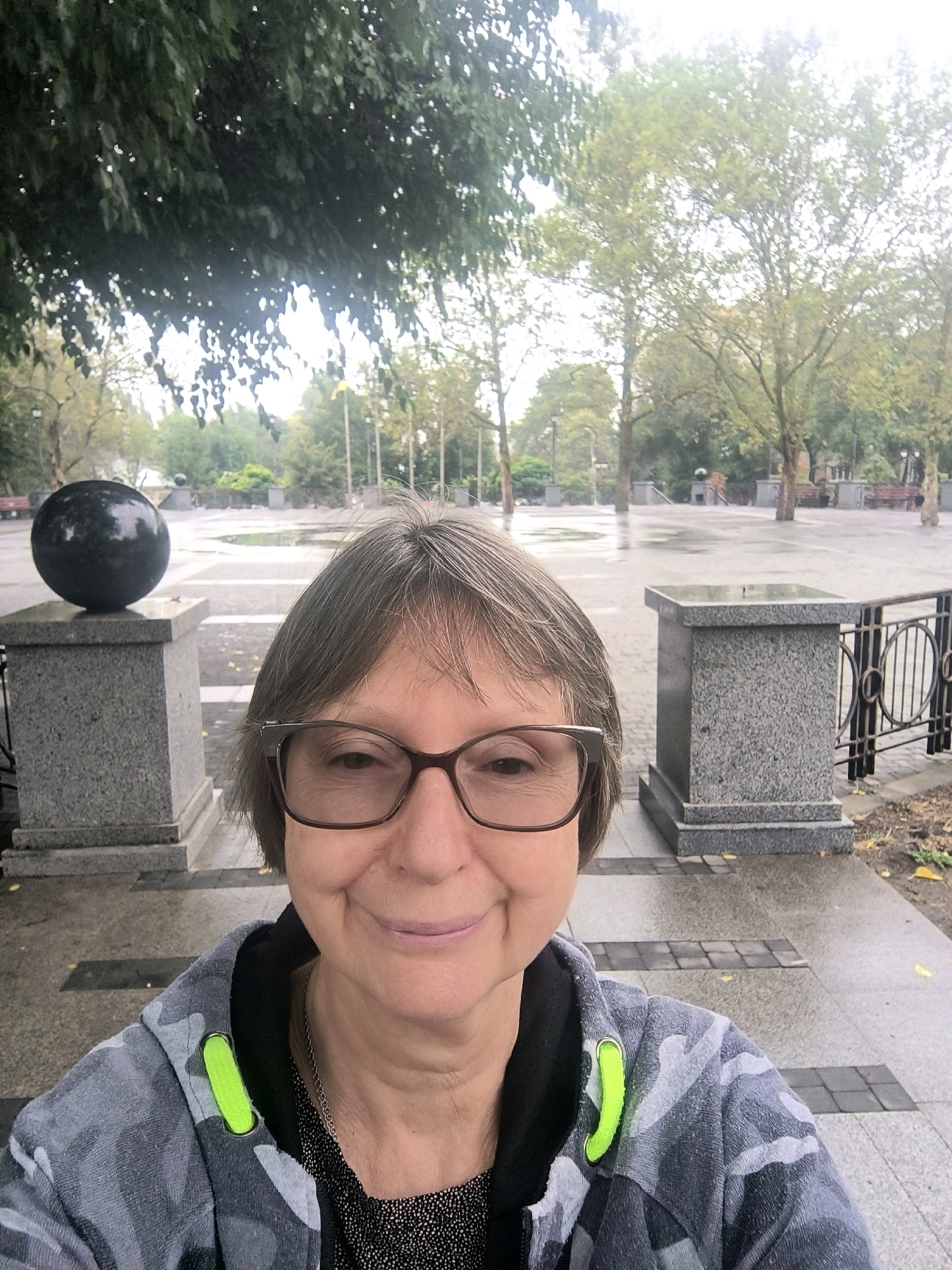 A woman with short, graying hair and glasses is taking a selfie in an outdoor, paved plaza that appears wet from rain. She is wearing a camouflage-patterned hooded sweatshirt with bright green zipper pulls and a dark, speckled shirt underneath, along with a silver necklace.
The plaza has a modern, minimalist design with several large, granite pedestals. One pedestal on the left holds a large, black, spherical sculpture. Another pedestal is visible on the right, with a black metal railing featuring circular designs. The ground is paved with rectangular stones, and there are puddles indicating recent rainfall. Trees with sparse foliage are scattered throughout the background, and benches can be seen in the distance. The overall atmosphere is muted and somewhat overcast.
The scene is located in Mykolaiv, Ukraine, likely within a public park or square. The presence of the unique spherical sculpture and the architectural style of the pedestals suggest a specific landmark or public art installation within the city.