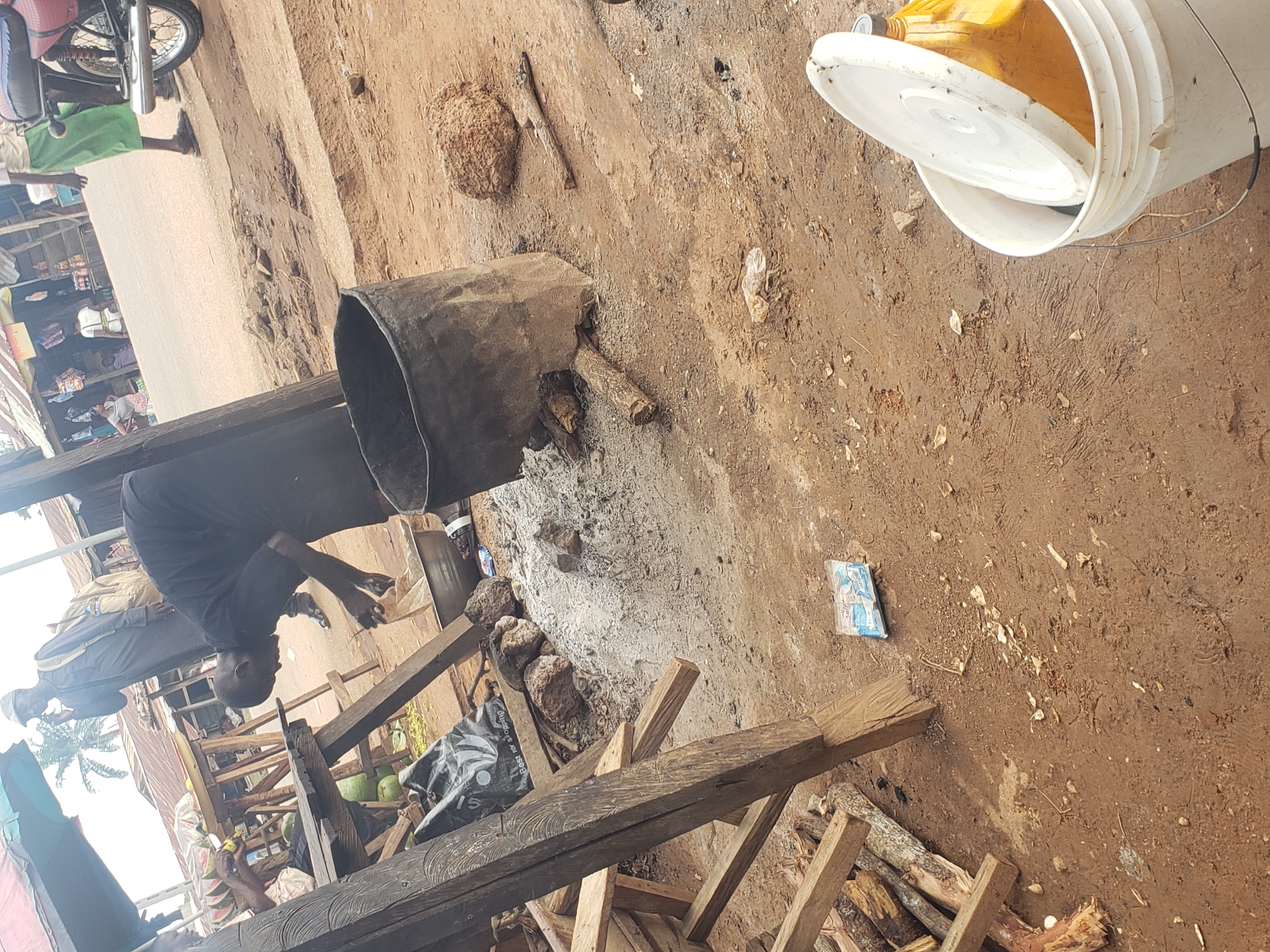 In a street market setting in Kachia, Nigeria, a person in a black garment is seen tending to a metal pot filled with a grey substance. Beside the pot, several stones and a piece of wood are scattered on the dusty ground. In the foreground, a white bucket with a lid is positioned, containing a yellow liquid. To the left, stacks of firewood are visible, along with a wooden structure that appears to be a stall. In the background, a motorcycle and other market stalls with goods are present, and people are engaged in activities related to the market. The overall environment is outdoor and appears to be a busy marketplace.
