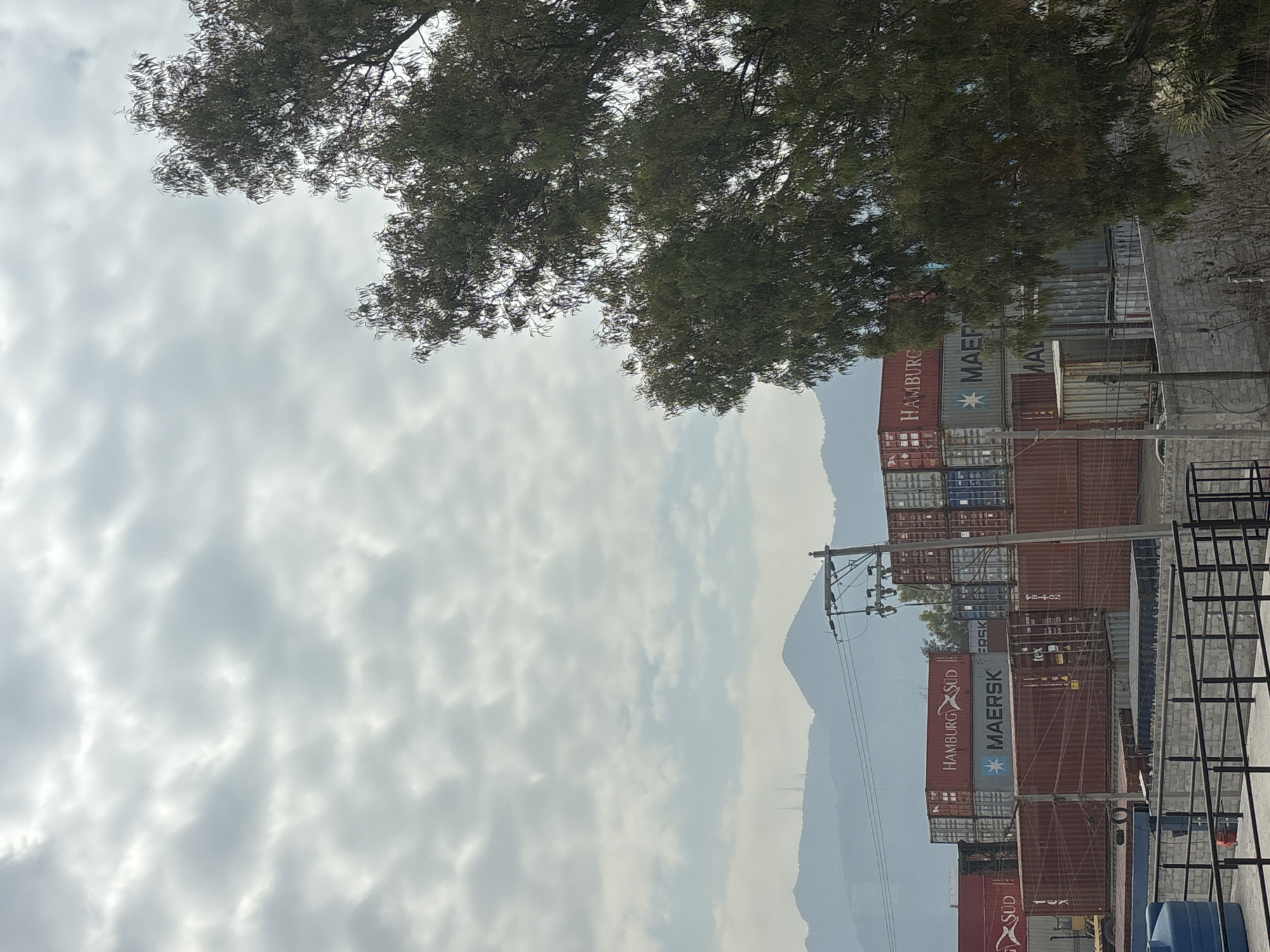 The image depicts a landscape with a view of stacks of shipping containers near Buenavista, Mexico. In the background, there's a hazy mountain range under a cloudy sky. The foreground includes a power line and some metal structures. A tree occupies the top portion of the frame, partially obscuring the view. The containers are labeled with the names "Hamburg Sud" and "Maersk".