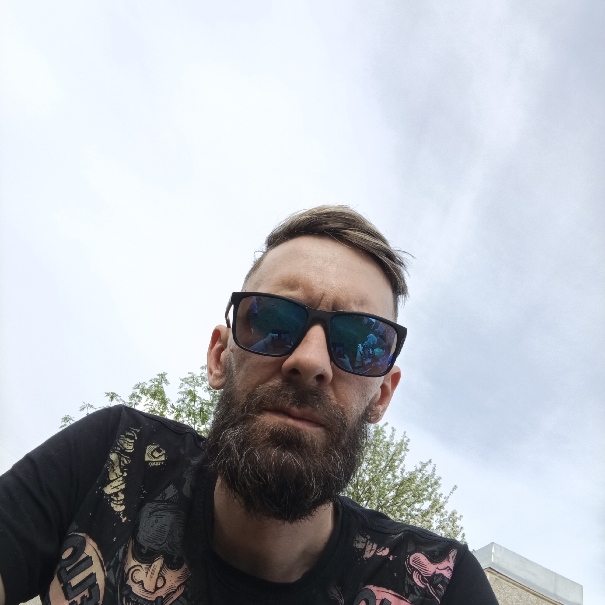 A close-up, low-angle photograph shows a man with a beard wearing sunglasses outdoors. The man has short brown hair and is wearing a black t-shirt with a faded graphic print. His sunglasses reflect a crowd of people, some holding up phones, suggesting an event or performance. The background features an overcast sky and trees with green leaves. The lower right corner of the image includes a portion of a building with a light-colored exterior and a gray roof. Based on the request it may be in Марковский, Russia.