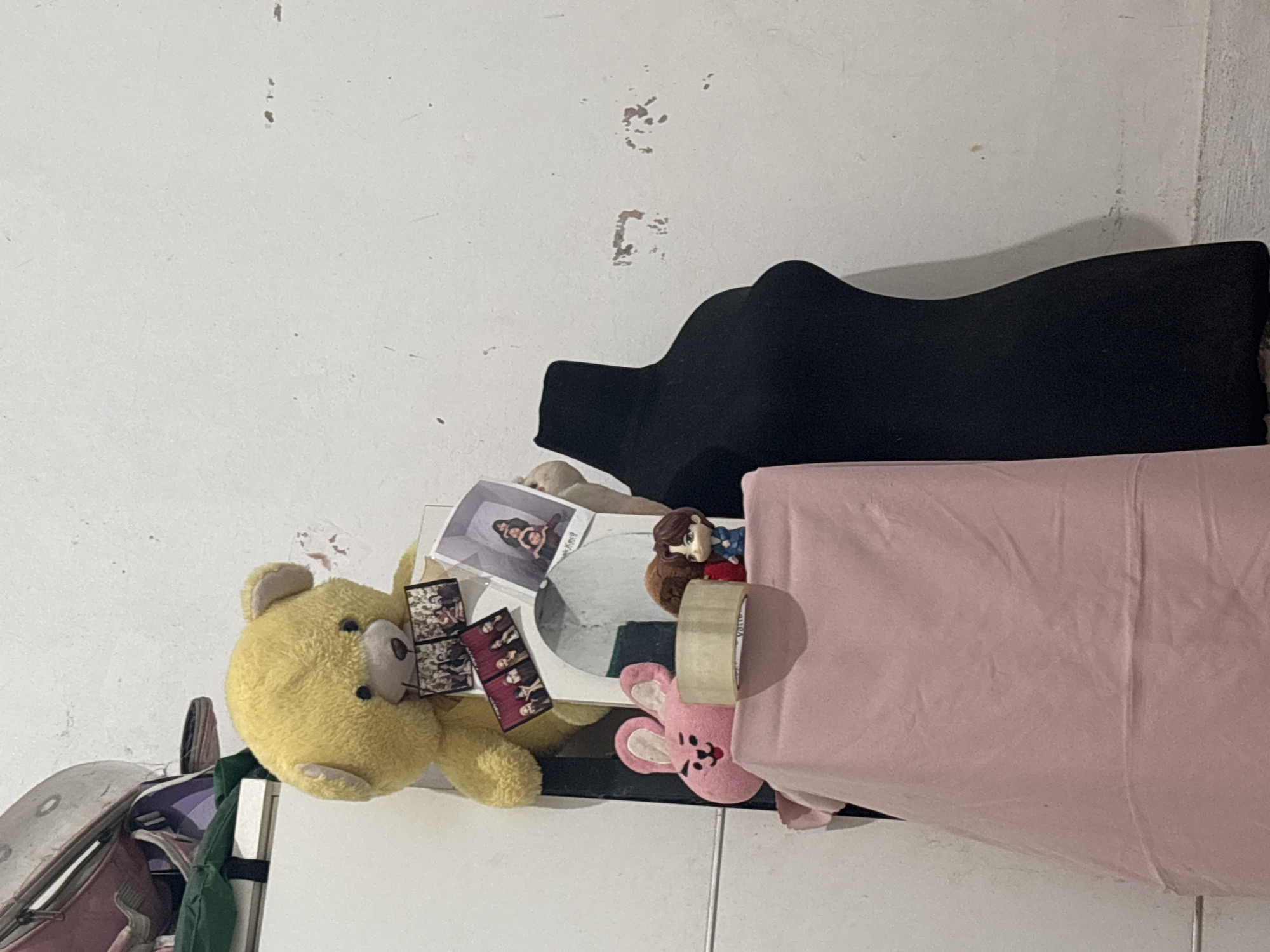 In North Jakarta, Indonesia, a stuffed yellow teddy bear is positioned on a shelf, holding several small photographs. To its left, a pink plush rabbit with long ears is visible. In front of the bear and rabbit, a roll of tape sits on a draped pink fabric. Behind these items, a black mannequin torso stands to the right. The wall behind the objects is painted white and shows signs of wear, with peeling paint and small marks. The lighting is dim, casting shadows on the wall.