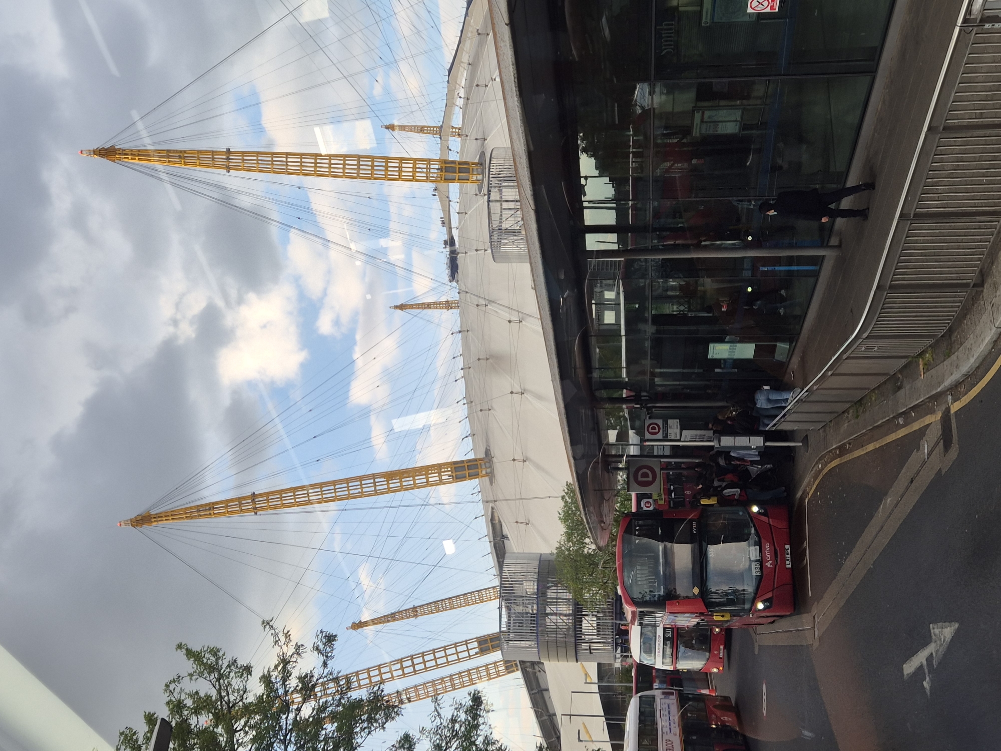The image shows the O2 Arena in London, United Kingdom. The arena's iconic dome structure with its support masts is visible against a sky with clouds. To the right of the arena, there is a building with glass windows reflecting the surrounding environment. In front of the building, a queue of people waits at a bus stop where several red London double-decker buses are lined up. The road is marked with yellow lines and a painted arrow. A tree is visible in the lower left corner of the image. A person wearing black is standing beside the building's glass window.