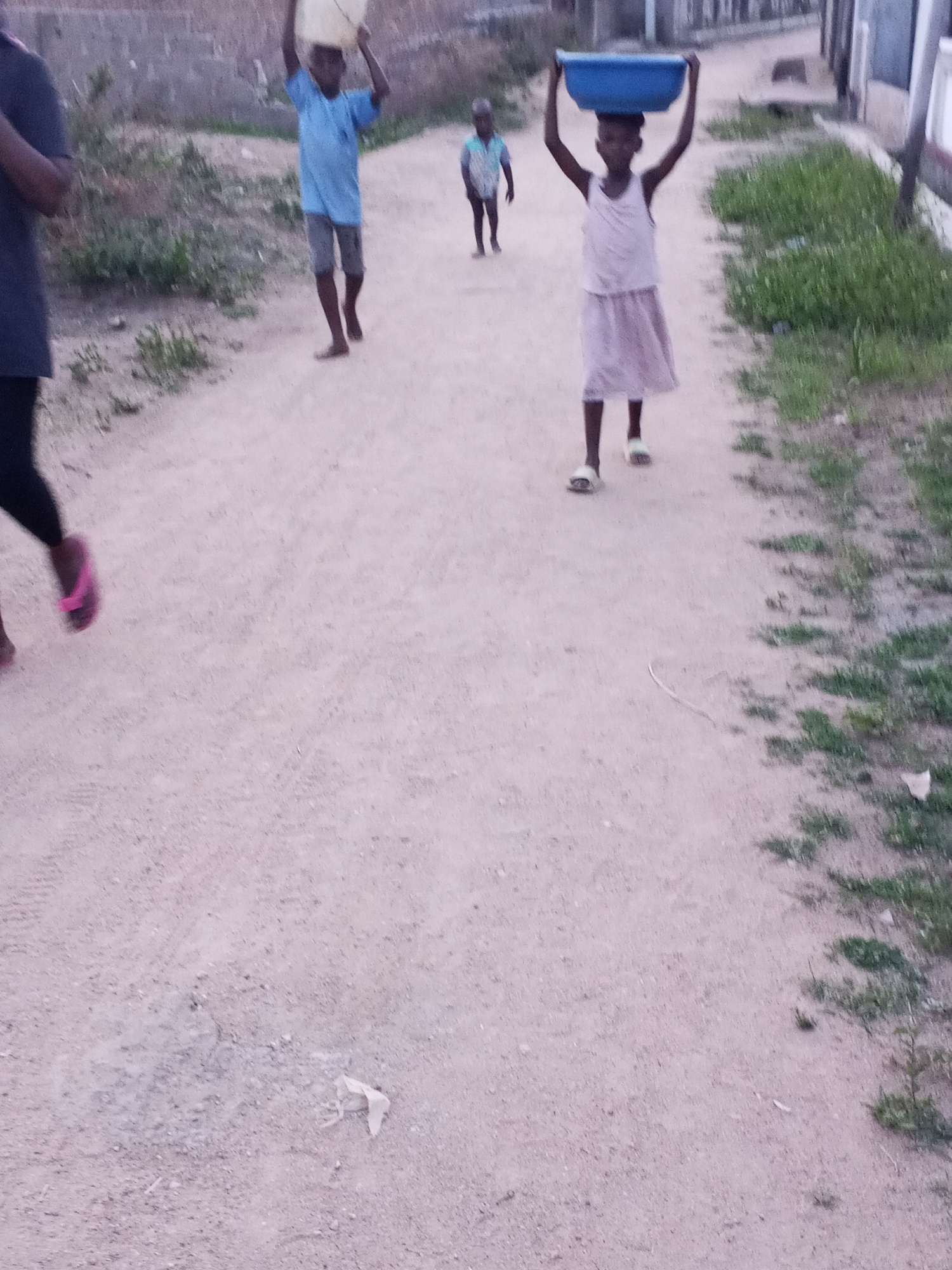 The image shows four people on a dirt road in Zango-ndaji, Nigeria. The road is flanked by sparse vegetation and some buildings. A girl in a light-colored dress and sandals carries a blue basin on her head. A boy in a blue shirt and shorts carries a yellow container on his head. Another person wearing a dark shirt and shorts is walking behind him, partially obscured. Another person, in a striped shirt, is walking further behind.