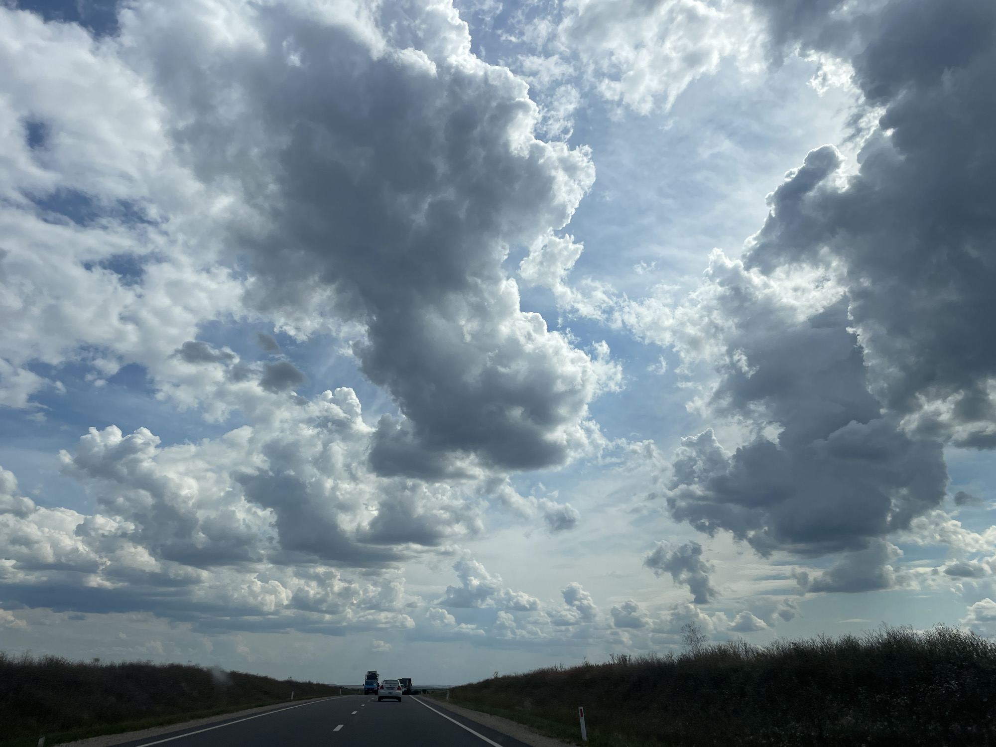 The image shows a view of the sky and a road, taken from inside a vehicle. The sky is filled with dramatic, large, grey and white clouds, with patches of blue visible in between. The road stretches into the distance and appears to be a highway. There are a few vehicles visible on the road, including at least one truck and one car, all moving away from the viewer. The sides of the road are lined with dark green vegetation and some sparse trees. The scene appears to be set in a rural or semi-rural area, given the open landscape and vegetation. While specific landmarks for Comrat, Moldova, are not identifiable in this generic highway scene, the cloudy sky and the type of vegetation are consistent with what might be found in the Moldovan countryside.