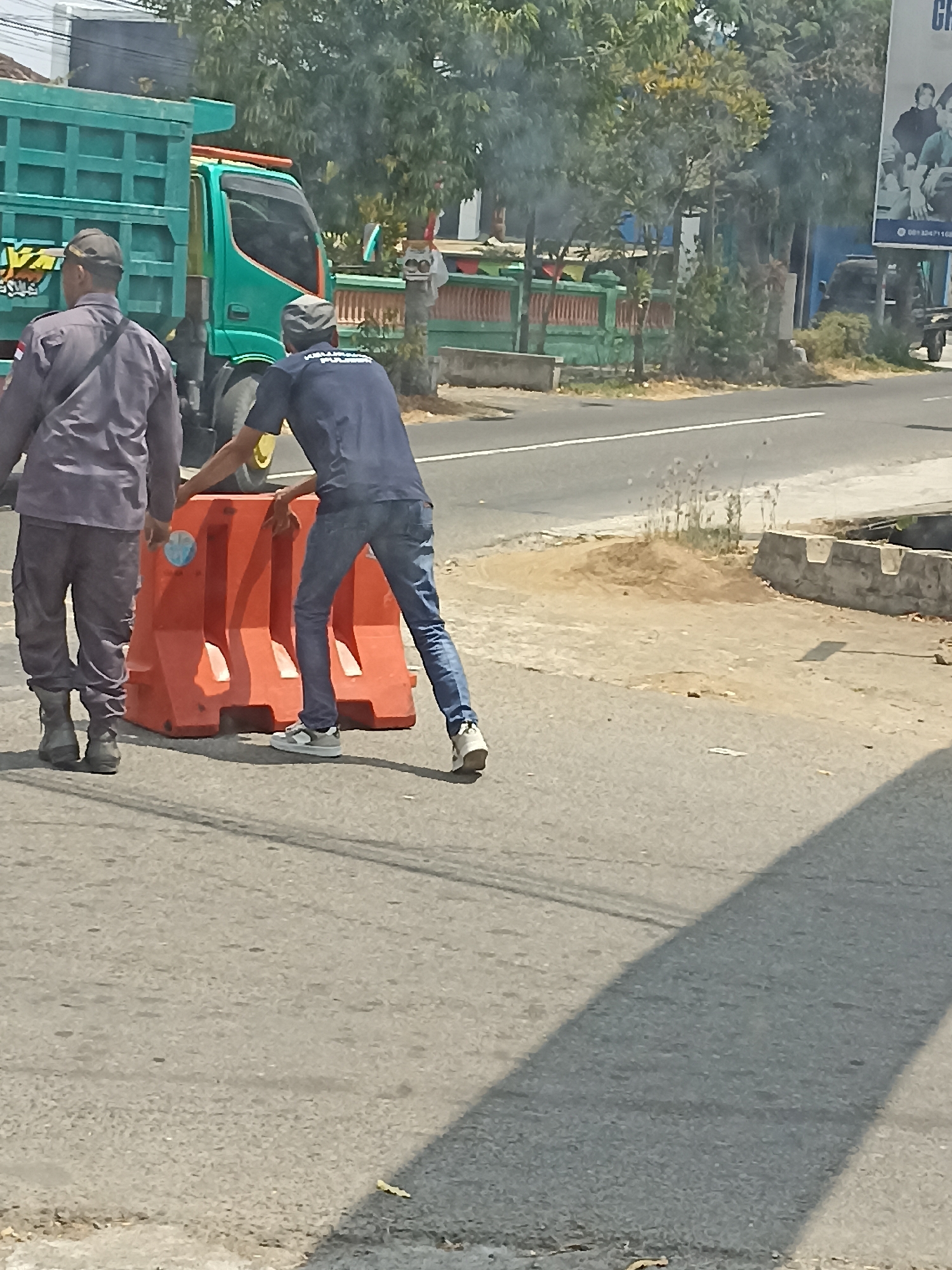 In the city of Pulisen, Indonesia, two men are in the process of moving an orange traffic barrier on a paved road. The man on the left is wearing a dark shirt and pants with a cap, while the man on the right is wearing a blue shirt, jeans, and sneakers, pushing the barrier. A green truck is parked on the right side of the road behind them. The environment appears to be a roadside with trees and a fence visible in the background.