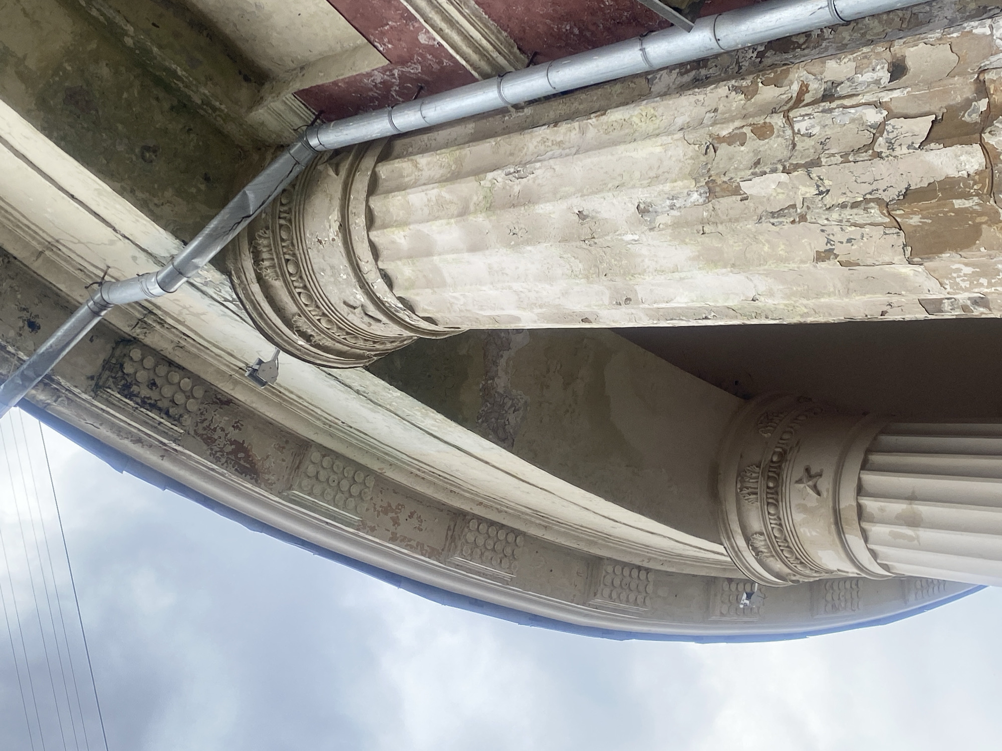 The image presents a detailed, low-angle view looking upwards at the weathered architectural elements of a grand building, likely in Moscow, Russia, against an overcast sky.
The primary subjects are classical architectural features: two large, fluted columns, their ornate capitals, and the elaborate cornice or entablature above them. The columns exhibit vertical grooves (fluting), and one capital is distinctly adorned with circular motifs and a star emblem, suggesting a classical or neoclassical style. The underside of the cornice (soffit) features decorative panels with repetitive circular patterns resembling dentils.
A notable detail is the extensive wear and tear on the building's surface. Paint is severely peeling, cracked, and discolored across the columns, capitals, and cornices, revealing darker, weathered layers underneath. This deterioration suggests significant age and exposure to the elements, hinting at a need for restoration or a long history.
Contrasting with the historic architecture is a modern-looking, grey metallic conduit or pipe running horizontally across the scene, secured by multiple clips to the underside of the structure. It shows a joint or bend, indicating its functional purpose, possibly for electrical wiring or drainage.
No people or specific activities are visible. The sky, seen at the bottom of the image, is overcast, suggesting a cloudy daytime setting. No visible text is present on the building's facade or the conduit. The overall impression is one of weathered grandeur, where historic beauty coexists with signs of aging and modern utility installations.